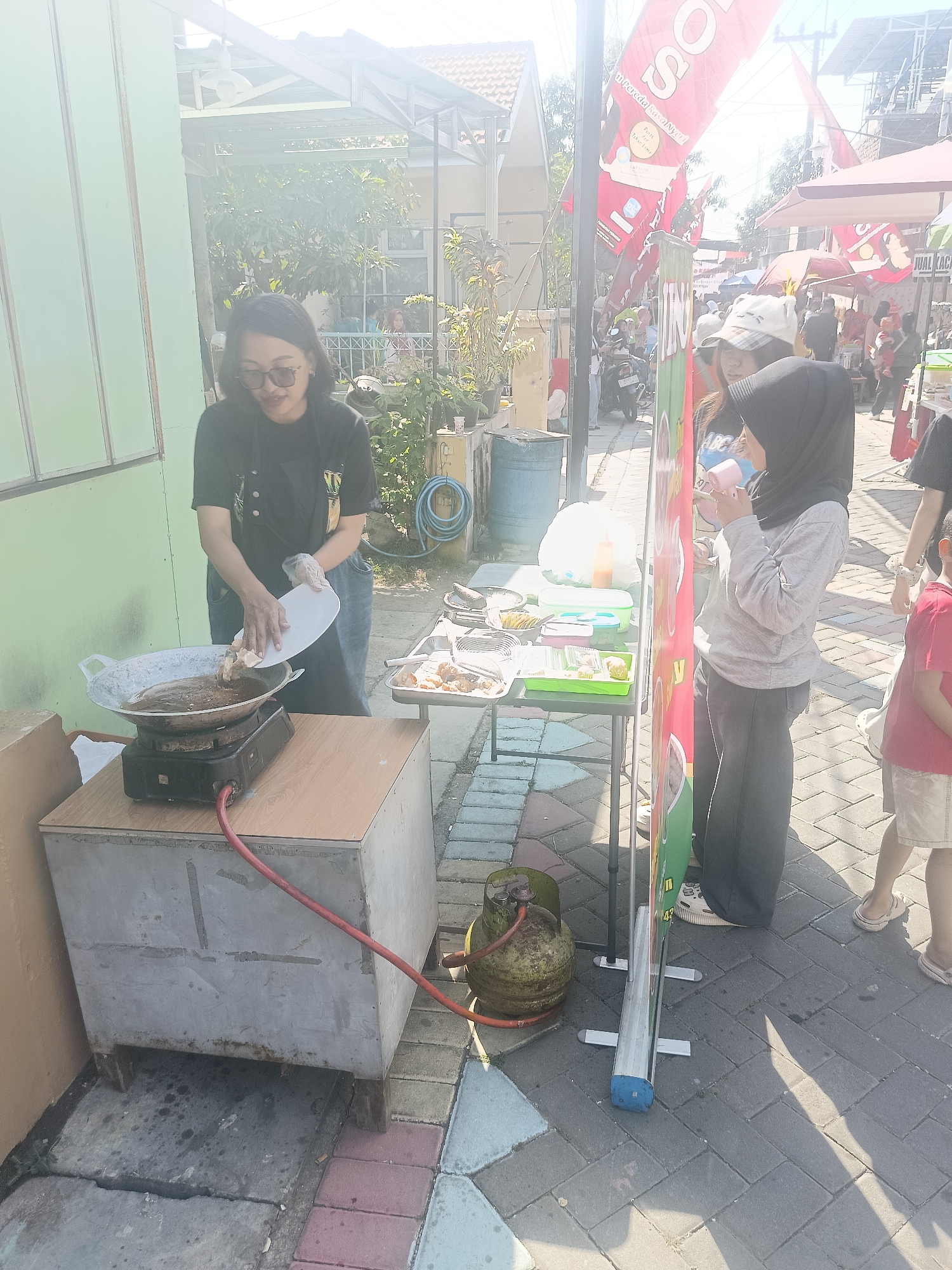 A woman in a black t-shirt and jeans, wearing glasses and gloves, is frying food in a wok placed on a portable stove. The stove is connected to a green gas tank with a red hose. She is holding a white plate with food, about to put it into the hot oil. The scene appears to be an outdoor food stall or market, likely in Gedangan, Indonesia, as suggested by the presence of red and white banners commonly seen at Indonesian events and shops. In the background, other people, stalls, and buildings are visible, under bright sunlight. The ground is paved with interlocking blocks. A younger person in a gray hijab is standing next to a colorful display banner.