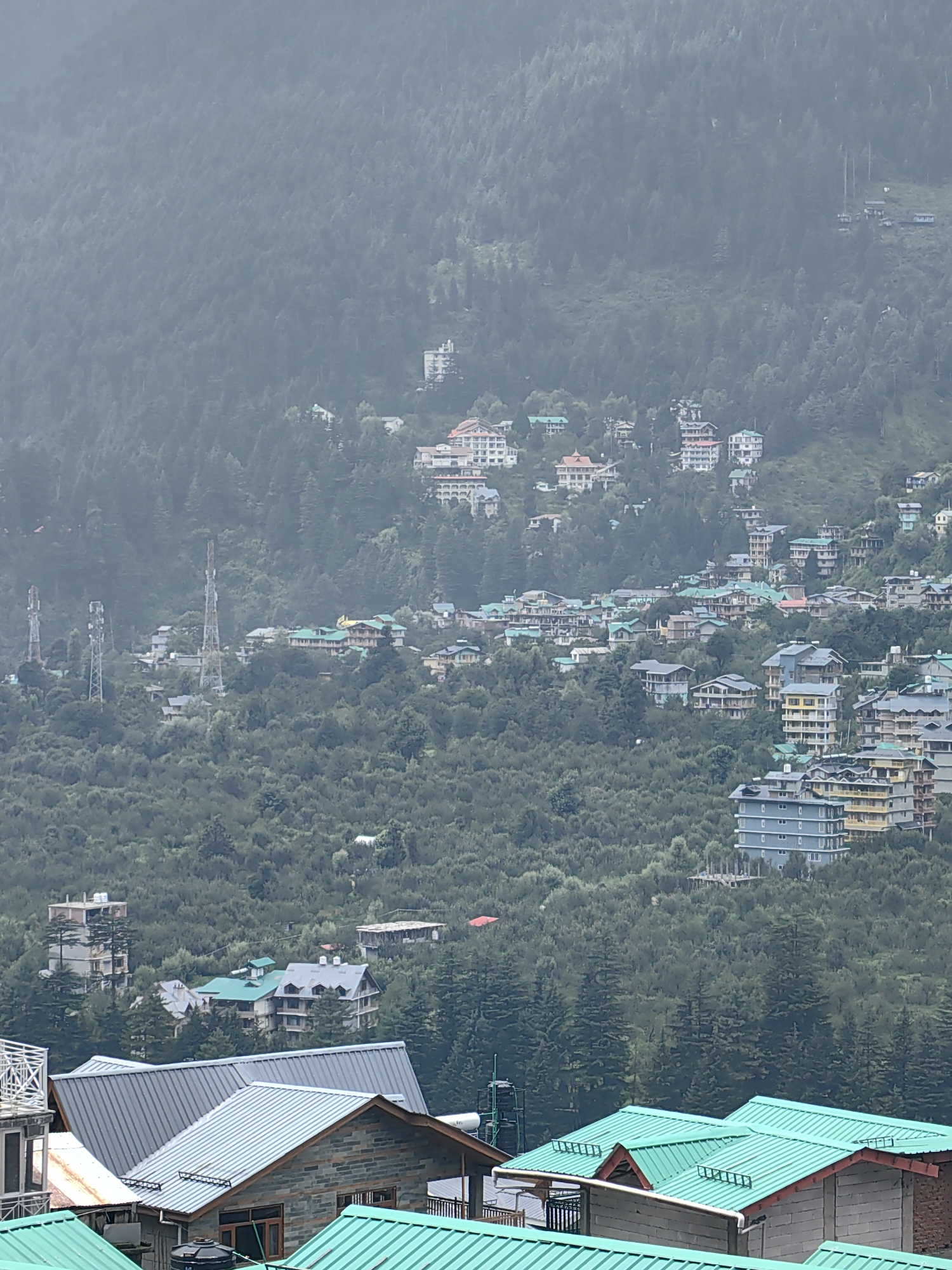 The image depicts a hillside view of Vashisht, India, characterized by a dense forest covering the slopes and numerous buildings scattered throughout. The foreground features several structures with distinctive green corrugated metal roofs, some appearing to be residential or commercial buildings. Further up the hill, a collection of multi-story buildings, hotels, and houses, many with white or light-colored facades and tiled or flat roofs, are visible, blending into the natural landscape. Communication towers are also present on the hillside. The atmosphere appears hazy or overcast, suggesting cloudy weather or atmospheric conditions common in mountainous regions. There are no people or active interactions visible in the scene.