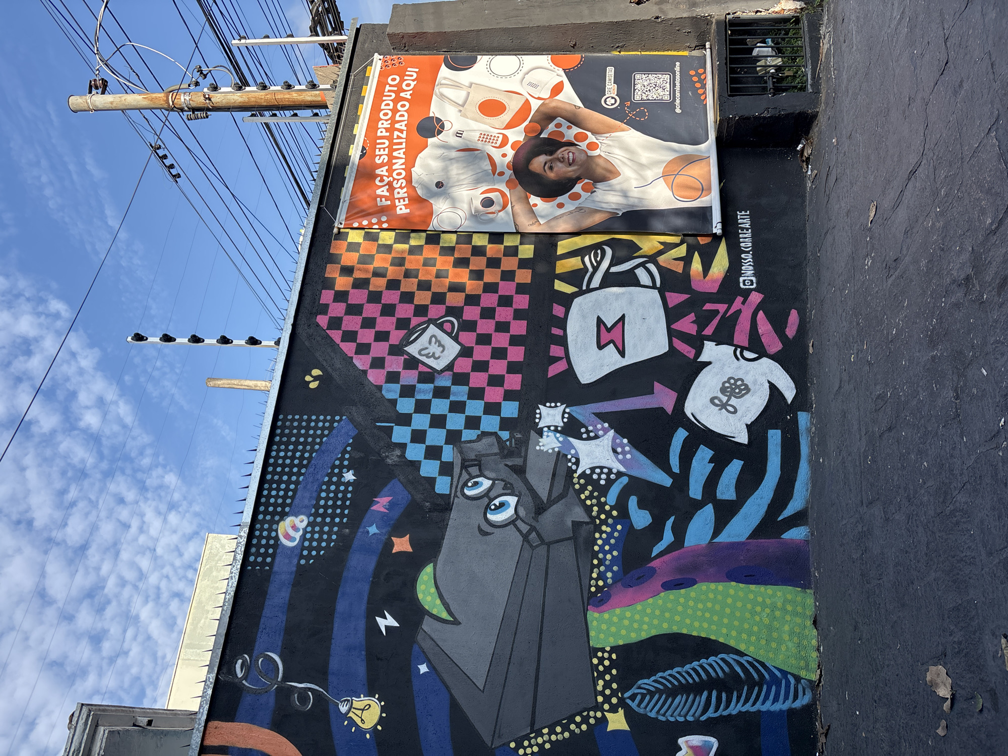 This vertical outdoor shot captures a vibrant street art mural on a dark building wall, topped by a promotional banner, set against a bright blue sky. The scene is characteristic of an urban environment, identified as São Paulo, Brazil.
The rectangular banner features a smiling young person with dark curly hair and a hat, wearing a white t-shirt, depicted lying down. They are surrounded by various customizable products such as tote bags, mugs, caps, and phone cases, presented in white with orange circular designs. Text on the banner reads "FAÇA SEU PRODUTO PERSONALIZADO AQUI" (Make your personalized product here), alongside a QR code and the social media handle "@criecamisetasonline."
Below the banner, the colorful mural unfolds with a mix of abstract and graphic elements on a black background. A central, grey, geometric figure with three eyes is prominent. Other designs include a pink and black checkerboard pattern, a white coffee mug with a leaf motif, a white tote bag with a lightning bolt, and a white teapot-like object with a flower. Scattered stars, lightning bolts, dots, and arrows animate the space, along with abstract blue, green, and purple shapes. A lightbulb hangs from a wire in the bottom left corner. The social media handle "@NOSSA.CORRE.ARTE" is painted in white on the wall, likely crediting the mural artists.
The sky is a clear, bright blue with a few scattered white clouds, suggesting a sunny day. A large, rusty utility pole with numerous wires runs vertically along the left side of the frame. Metal anti-climb spikes are visible along the top edge of the wall. A small metal grille can be seen above the banner on the upper right.