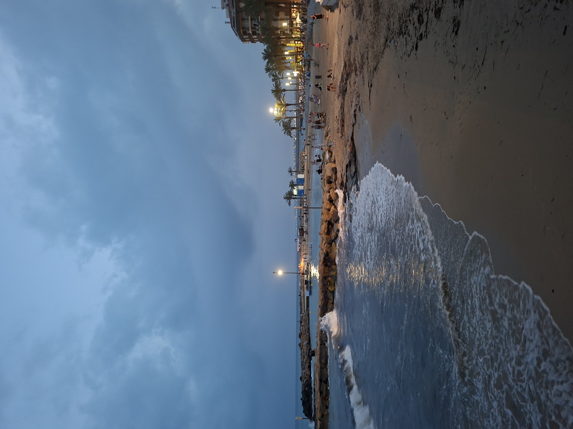 The image depicts a view of a beach at dusk in Torrevieja, Spain. The sky is overcast and a deep blue, with the clouds appearing heavy. The water is a dark blue-gray, with small waves gently lapping at the sandy shore and breaking against a rocky breakwater. Artificial lights are visible along the breakwater and the beachfront, illuminating the scene with a warm glow.
On the beach, there are scattered groups of people, some walking, some sitting or lying down, and others engaged in conversations. Palm trees are sparsely planted along the back of the beach, and a multi-story building with illuminated windows is visible on the right side of the frame, suggesting a developed area. The overall atmosphere is one of a calm evening, with people enjoying the beach as daylight fades.