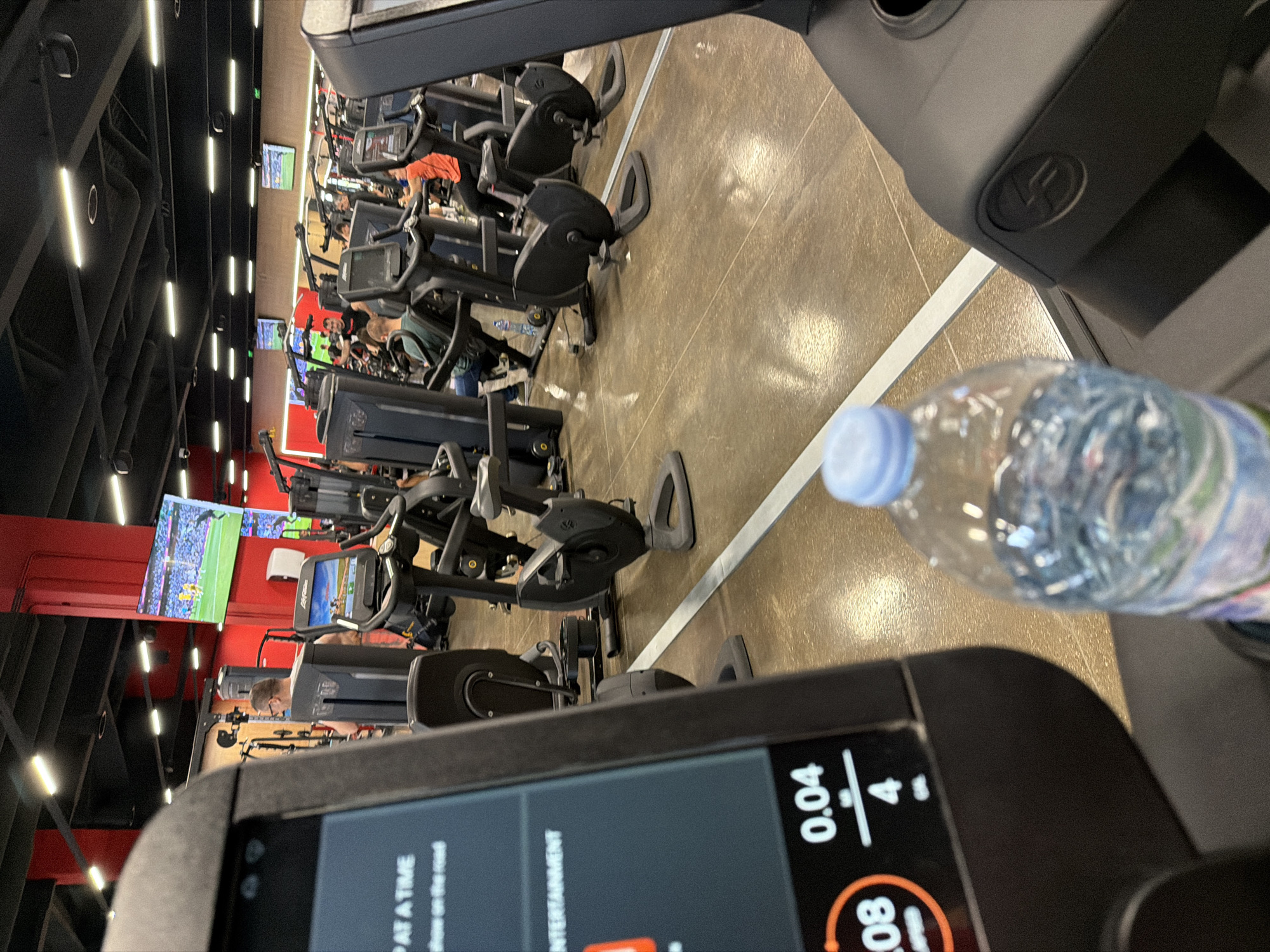 This image captures an indoor scene at a modern fitness center or gym in Plovdiv, Bulgaria, taken from the perspective of someone on an exercise machine. The photo is oriented vertically, with the view looking forward and slightly to the right, showing rows of cardio equipment and other gym amenities.
In the immediate foreground, a clear plastic water bottle with a light blue cap is visible on the right. Below and to the left, the screen of the exercise machine the photographer is using is partially visible, displaying digital information. Text on the screen includes "0.04" and "4" (likely representing distance and incline/level), an orange circle with "80" inside (possibly heart rate or speed), and phrases like "AT A TIME", "on the road", and "ENTERTAINMENT".
The midground features numerous exercise machines, including treadmills, elliptical trainers, and stationary bikes, arranged in organized rows on a light brown, possibly polished concrete floor with a distinct white dividing line running through it. Several individuals are present, actively engaged in their workouts on various machines.
In the background, large flat-screen televisions are mounted on red walls, displaying what appears to be a live sports event, possibly a soccer or football match, given the green field and numerous players. The ceiling is dark with exposed structures and numerous bright, rectangular fluorescent light fixtures illuminating the space. A subtle "LF" logo (likely for Life Fitness) is visible on the console of the machine in the top right of the frame. The overall atmosphere suggests an active and well-lit gym environment, likely during the day or evening.