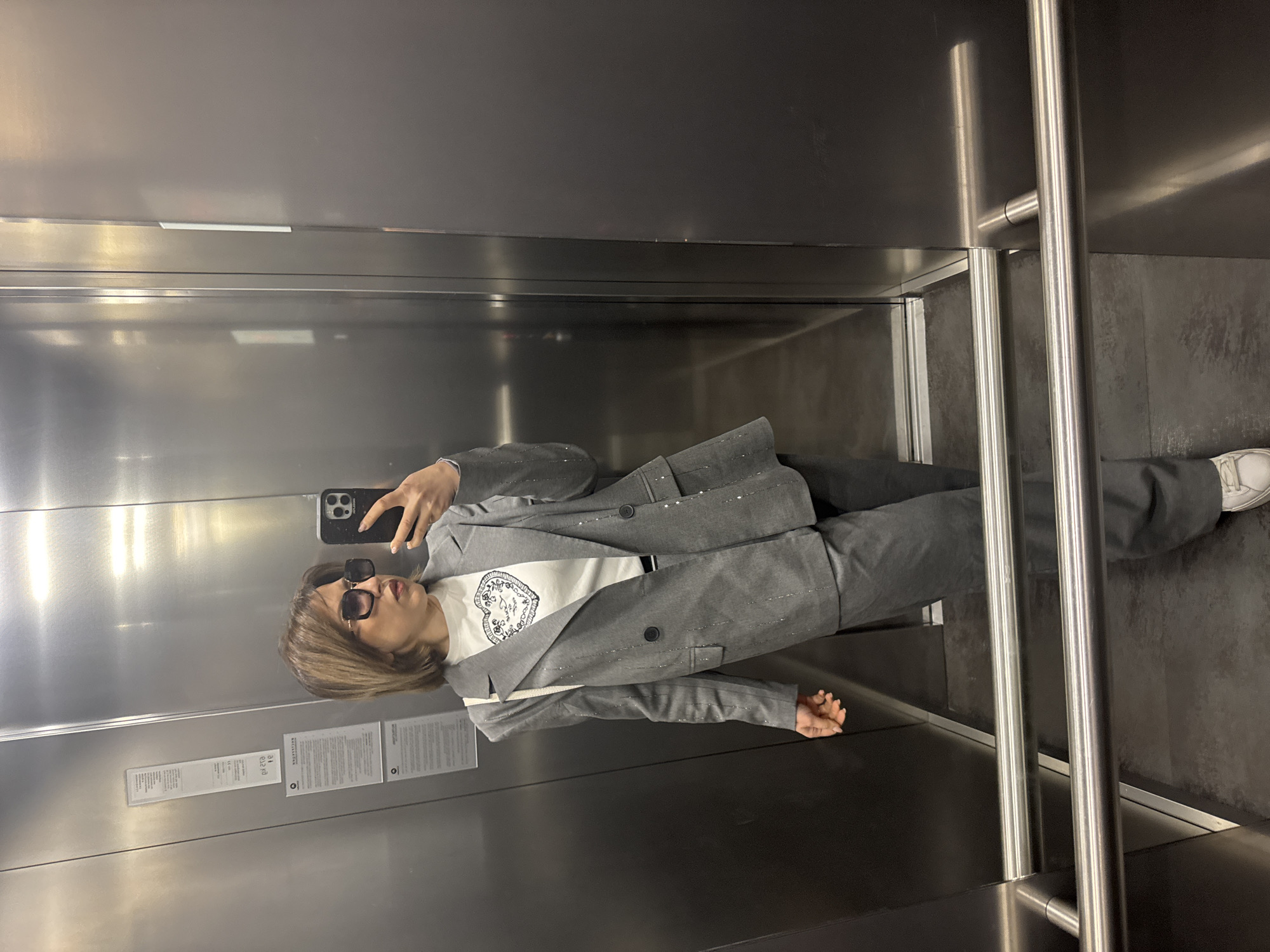 A woman in a gray suit with rhinestone embellishments and sunglasses stands in an elevator, taking a selfie with a smartphone. She wears a white t-shirt with a graphic design visible. The elevator has stainless steel walls and handrails, with safety and information notices posted inside. The floor appears dark gray. The location is likely in the city of Sofia, Bulgaria, but this cannot be definitively confirmed from the image alone.