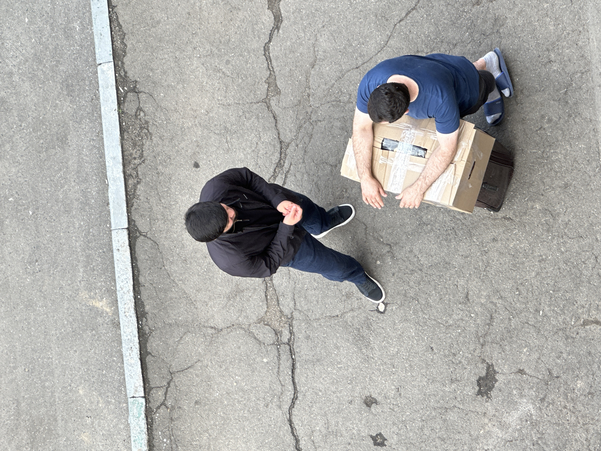 Two men are on a cracked asphalt surface, possibly in Podolsk, Russia, indicated by a curb with light blue paint on its edge. The man on the left, wearing a black jacket and blue jeans, stands with his hands clasped in front of him. The man on the right, in a blue t-shirt and dark shorts, is kneeling and handling a cardboard box. A dark suitcase is placed under the box. The scene appears to be outdoors, with the texture of the asphalt and cracks suggesting an urban environment.