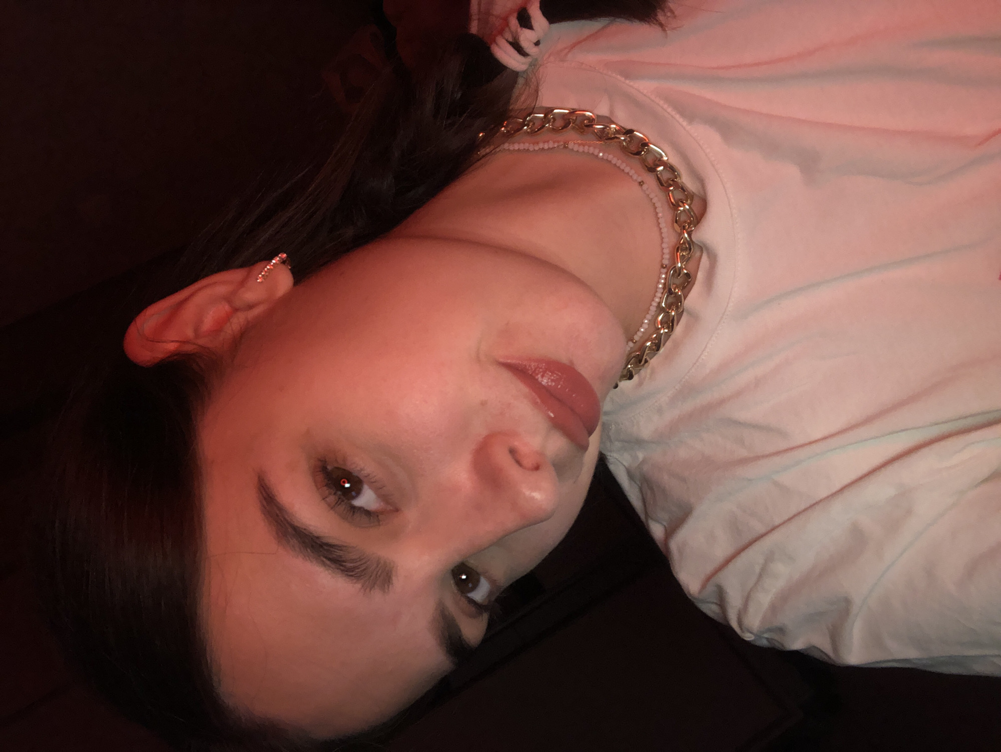 The image shows a close-up, head-and-shoulders shot of a young woman with dark hair and fair skin, illuminated by red lighting. She is wearing a white t-shirt and a gold chain necklace layered over a smaller beaded necklace. She has a small, sparkling earring in her visible left ear. Her gaze is directed slightly away from the camera, and her lips are slightly parted. The background is dark and indistinct. There is no information in the image to suggest a location in Noviy, Russia.