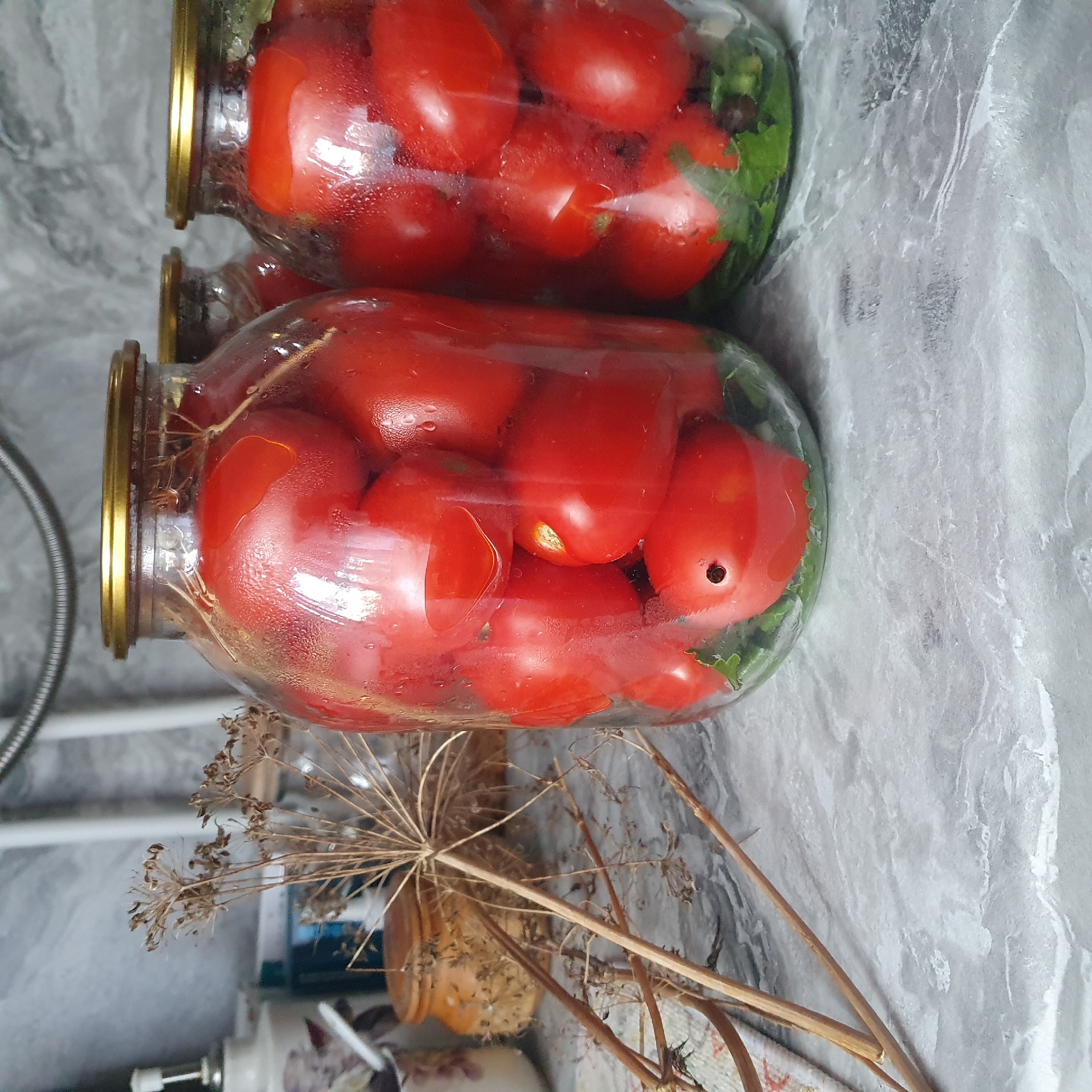 This media file features two glass jars filled with red tomatoes and green herbs, likely for preservation. The jars have gold-colored lids and are placed on a marble-patterned surface. In the foreground, dried dill stalks are visible, adding a rustic touch to the scene. The lighting suggests it might be daytime, and the overall setting appears to be a kitchen or pantry. There are no people visible in the image, and the focus is on the preserved food items. The image is oriented vertically.