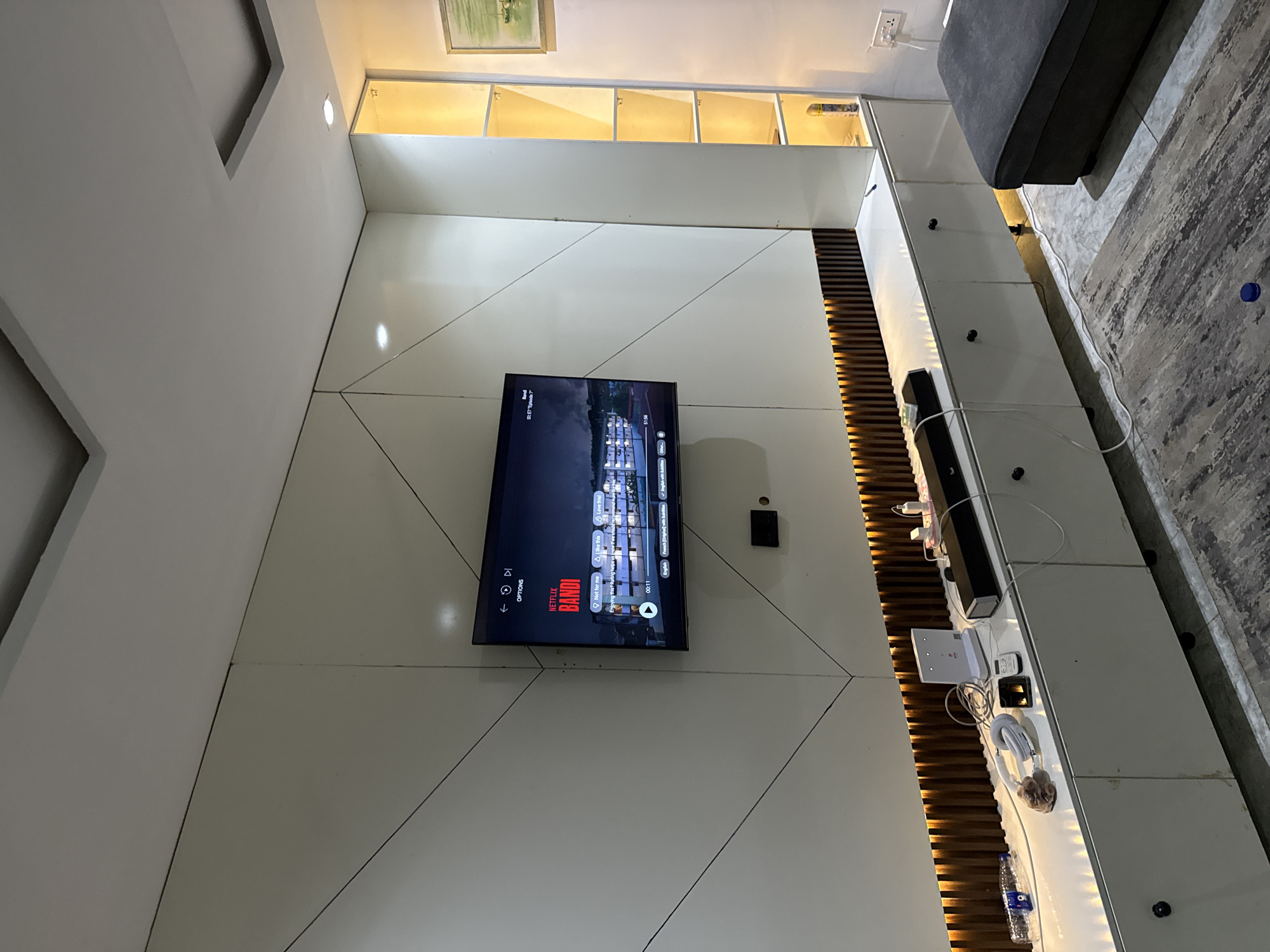 The image shows an interior view of a room, likely a living area. The focus is on a large television mounted on a geometric, white paneled wall. The TV is displaying a Netflix screen with a show titled "BANDI." Above the television are open shelves with a light-colored interior.
To the right of the television is a vertical wooden slat design and a low, white cabinet cluttered with various electronic devices and wires. Above the cabinet are knobs attached to the face. A section of a gray sofa is visible in the upper right corner. The room has overhead lighting recessed into the ceiling, and there's a rug on the floor next to the sofa. The location is not discernible as being in Iddo Tudunwada, Nigeria from the given image.