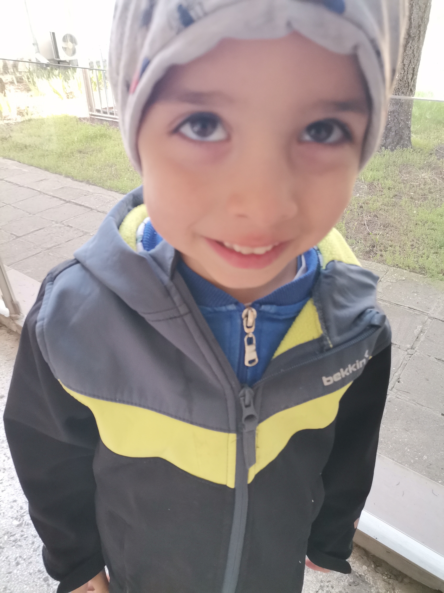 A young child, likely a boy, is the primary subject of the image. He is wearing a light grey beanie and a jacket with a dark grey upper part, a yellow horizontal stripe, and a black lower part. Underneath the jacket, a blue zippered top is visible. The child is smiling slightly, looking upwards towards the camera.
The setting appears to be outdoors on a paved walkway, possibly in a residential area. In the background, there is a section of a fence, a tree, and a building with an air conditioning unit mounted on the exterior. Based on visual context, the location is presumed to be within the city of Dobrich, Bulgaria.