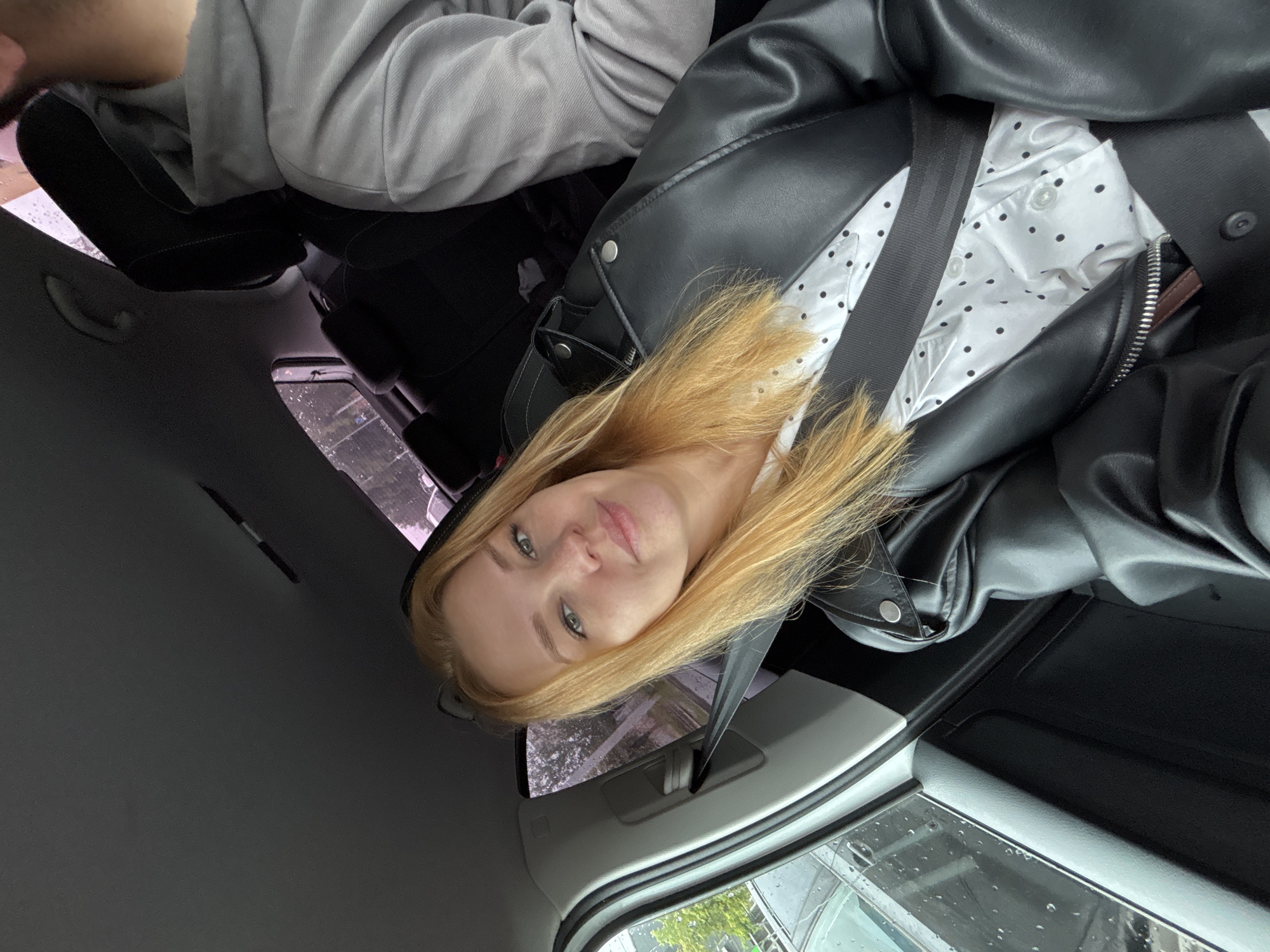 A blonde woman with fair skin and green eyes is captured in a selfie taken from inside a car. She is wearing a black leather jacket over a white shirt with black polka dots. A black seatbelt is fastened across her chest. The interior of the car is visible, including a dark-colored headrest and a grey hooded sweatshirt of another person in the passenger seat. Outside the car window, raindrops are visible on the glass, suggesting inclement weather. The background shows trees and possibly buildings, indicating an urban or suburban setting. The scene appears to be in the city of Zavolzhye, Russia.