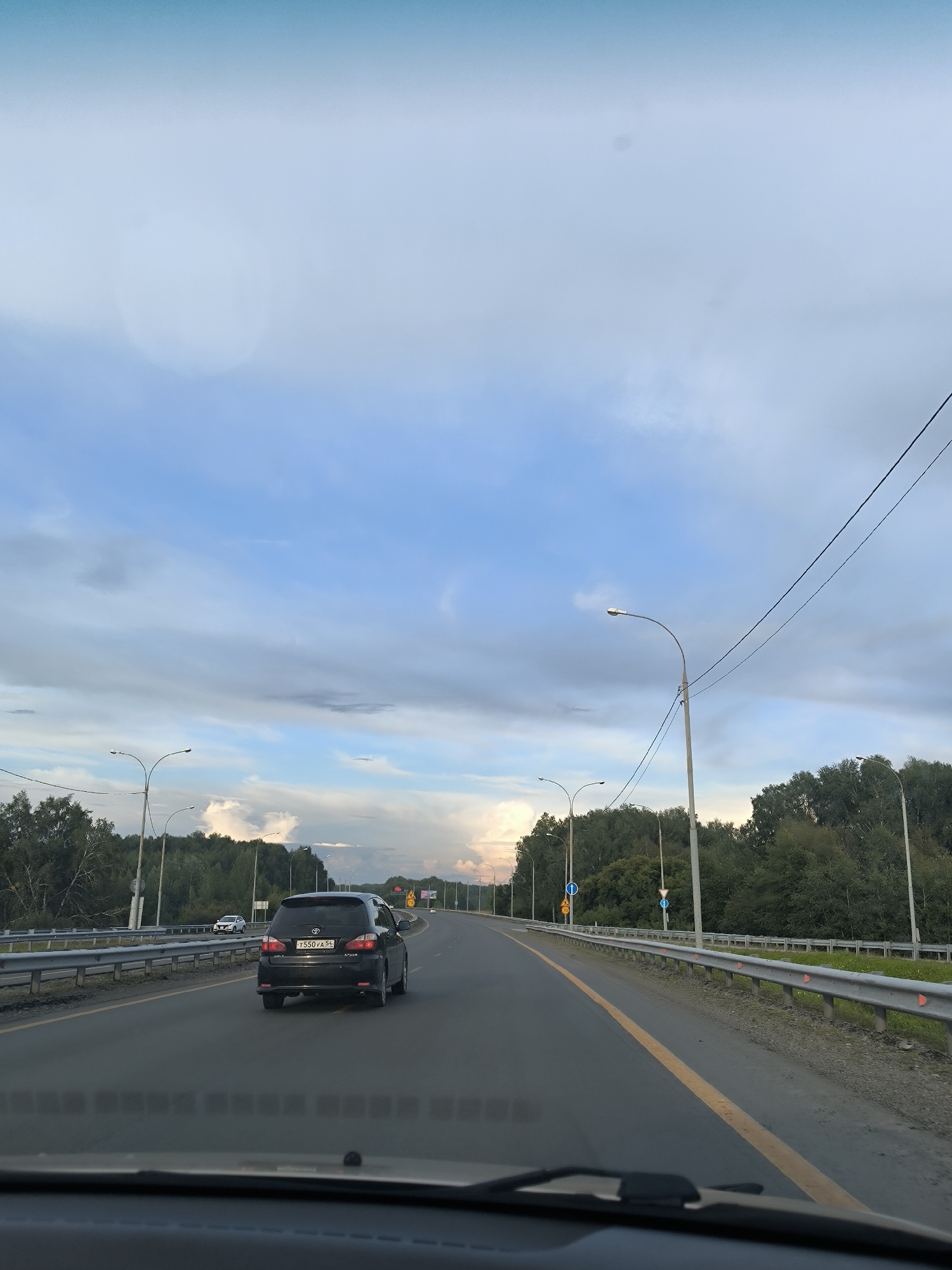 The image captures a view from inside a vehicle driving on a multi-lane highway on the outskirts of Novosibirsk, Russia. The road has a yellow dividing line and is bordered by metal guardrails. Streetlights are positioned along the roadside. Dense green trees line the right side of the highway. A black Toyota Ipsum car with the license plate T550VA 54 is in the foreground on the right lane. In the distance, there are other vehicles and more trees. The sky above is partly cloudy with shades of blue and gray, and there is a hint of sunset colors on the horizon.