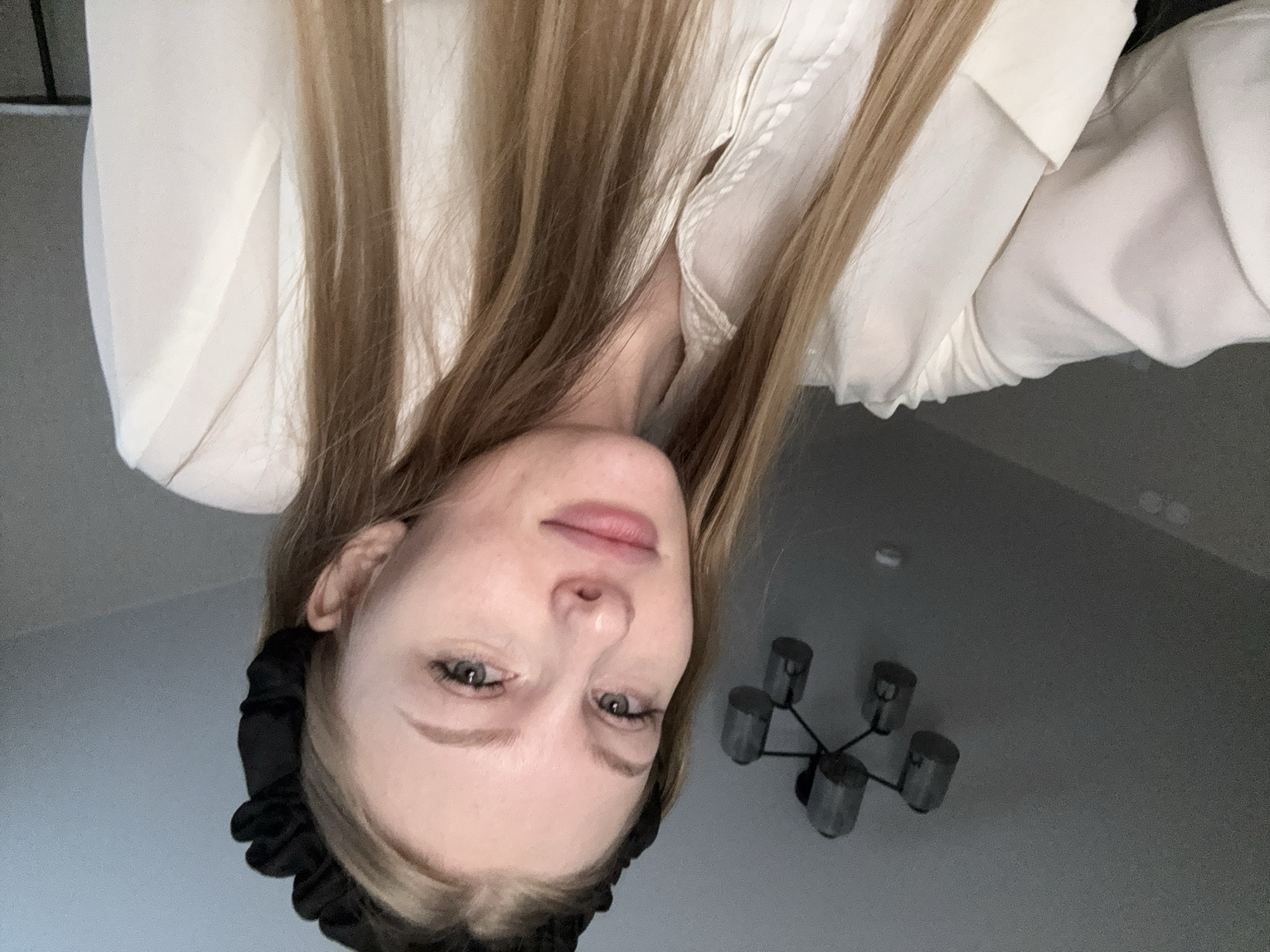 A young woman with blonde hair and a black scrunchie headband is pictured from the chest up, looking towards the camera. She is wearing a white collared shirt with buttons. The background is a plain wall with a dark ceiling fixture consisting of multiple cylindrical lights. The setting does not provide any specific visual indicators of its location within the city of Perm, Russia.