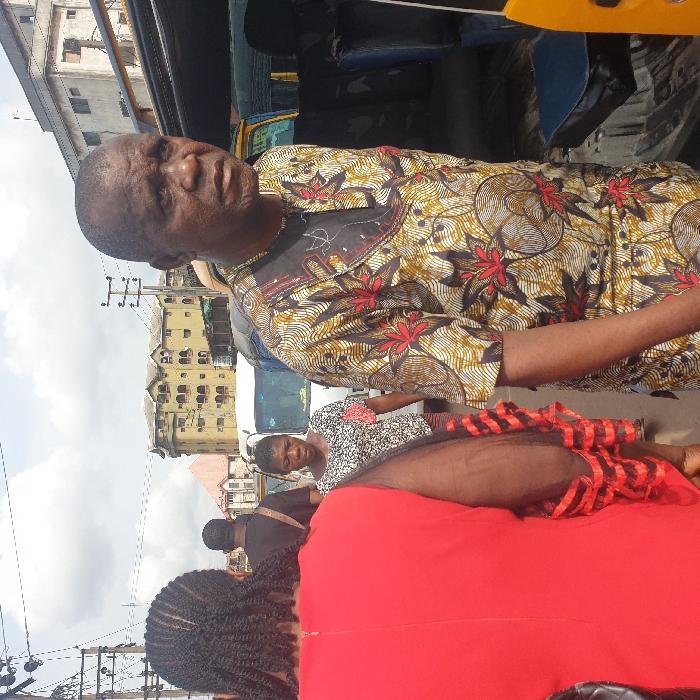 A daytime street scene in Onitsha, Nigeria, depicts an active urban environment with multiple pedestrians and vehicles. In the foreground, a bald adult male, appearing to be middle-aged, faces slightly right with a neutral to serious expression. He wears a short-sleeved traditional African patterned tunic in shades of brown, gold, red, and black, featuring floral and swirl motifs. To his left, the back of an individual in a bright red dress with dark braided hair is visible. Their right arm, covered by a black sheer sleeve with red horizontal trim, extends into the frame.
Further into the midground, an adult female with short, dark hair walks towards the viewer, wearing a patterned black and white top with a red floral detail pinned to the chest, and a black and red striped skirt. Behind her, the back of an adult male in a black shirt with a dark satchel slung across his body is seen.
The background features multiple white vans or minibuses parked or in motion. On the far right, part of a yellow commercial vehicle, possibly a Keke Napep or a taxi, is visible. Multi-story buildings, some appearing weathered and aged, line the street under an overcast sky. Overhead utility lines and power poles are present throughout the upper portion of the scene. The ground surface appears to be paved, typical of a busy city street.