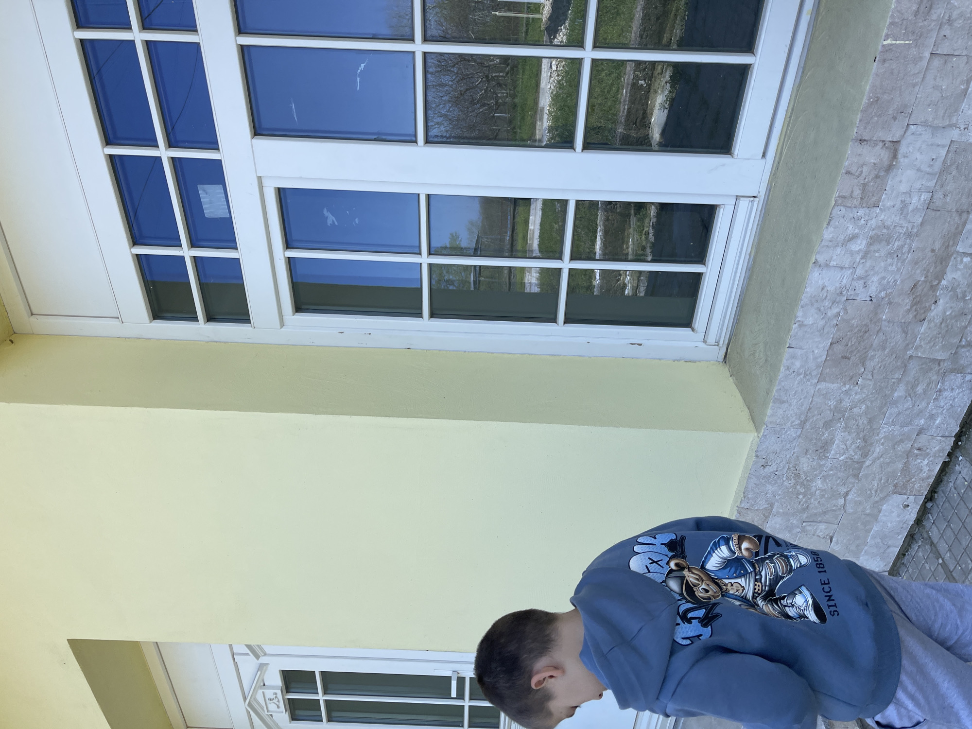 A low-angle, rotated, full outdoor shot depicts a young boy wearing a light blue hoodie and grey sweatpants standing in front of a pale yellow wall and white-framed windows. The back of his hoodie features a cartoon character and the words "SINCE 1850". The windows reflect an outdoor scene with grass, trees, and possible structures in the distance. The ground is paved with grey and white marble tiles. The setting appears to be in Somovit, Bulgaria.