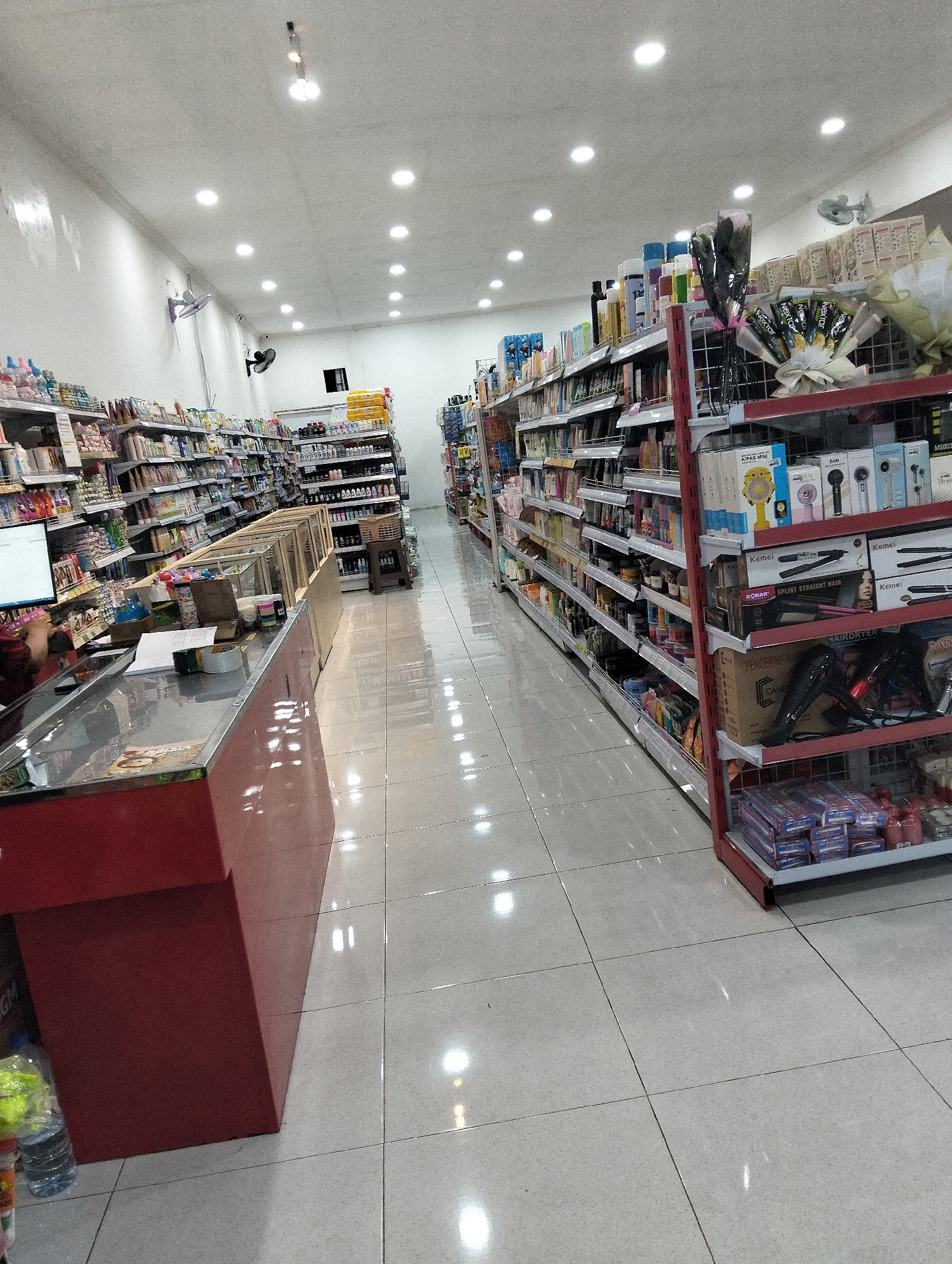 The image shows the interior of a brightly lit convenience store, likely in Kedungrejo, Indonesia. The store features tall shelves stocked with a variety of products, including toiletries, household goods, and packaged food items. A long counter is visible in the foreground, with a computer monitor and some papers on it. The floor is tiled and reflects the overhead lighting. Several ceiling fans are mounted on the walls and ceiling, and there are bright, circular lights illuminating the space. Some shelves display hair dryers and other personal care appliances. The overall impression is that of a well-organized and fully stocked retail environment.