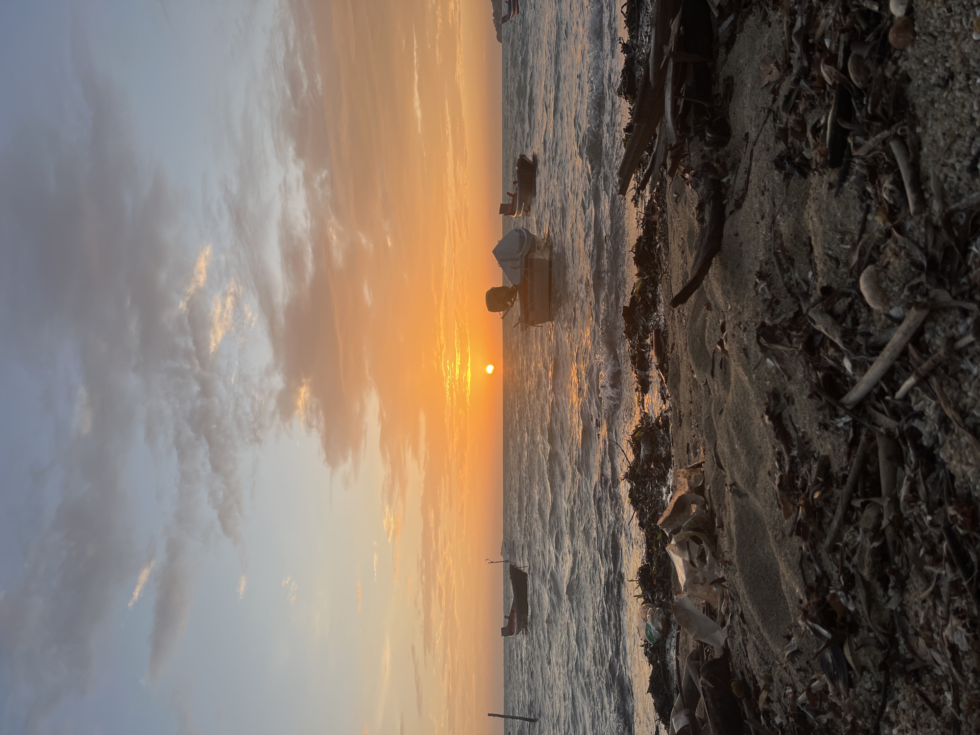 The image depicts a serene beach scene at sunset in the Ko Pha-ngan District of Thailand. The sun, a vibrant orange orb, is low on the horizon, casting a warm glow across the sky and water. Wispy clouds are scattered across the sky, tinged with shades of orange and pink from the setting sun. The sea is calm, with gentle waves lapping at the shore.
Several small fishing boats are anchored in the water, their silhouettes adding to the tranquil atmosphere. In the foreground, the beach is covered with sand, scattered with driftwood, shells, and some debris, including plastic bottles and packaging, suggesting a natural yet somewhat impacted environment. A single wooden pole stands in the water to the left of the frame.
The overall impression is one of peacefulness and natural beauty, juxtaposed with subtle signs of human presence and environmental impact. There are no people visible in the frame.