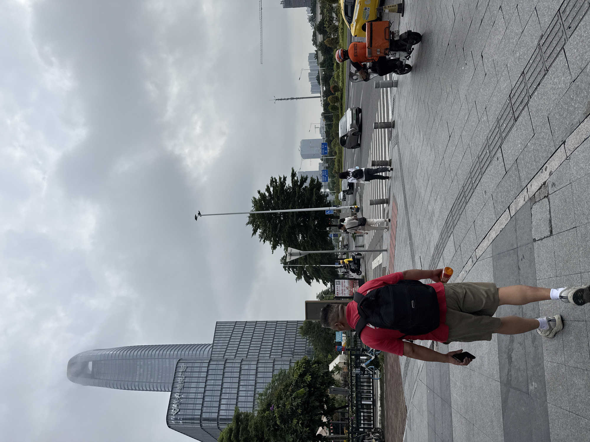 The image depicts a street scene in Haizhu District, China, under a cloudy sky. In the foreground, a man wearing a red t-shirt and khaki shorts walks away from the camera, carrying a black backpack and holding a bottle in his right hand and a mobile phone in his left. He is walking on a gray tiled sidewalk with tactile paving.
Further ahead, a pedestrian crossing is visible. Several people are crossing the street, including a woman with a backpack and another person in white. On the road, a scooter with an orange delivery box is being ridden by a person in an orange jacket. A yellow taxi is also visible.
The background features modern buildings, including a tall, curved skyscraper with a distinct top, and a multi-story glass-fronted building. Trees line the street, and there are streetlights and traffic signals. In the distance, more buildings and construction cranes suggest a developing urban area. The overall atmosphere is that of a typical city day with moderate activity.