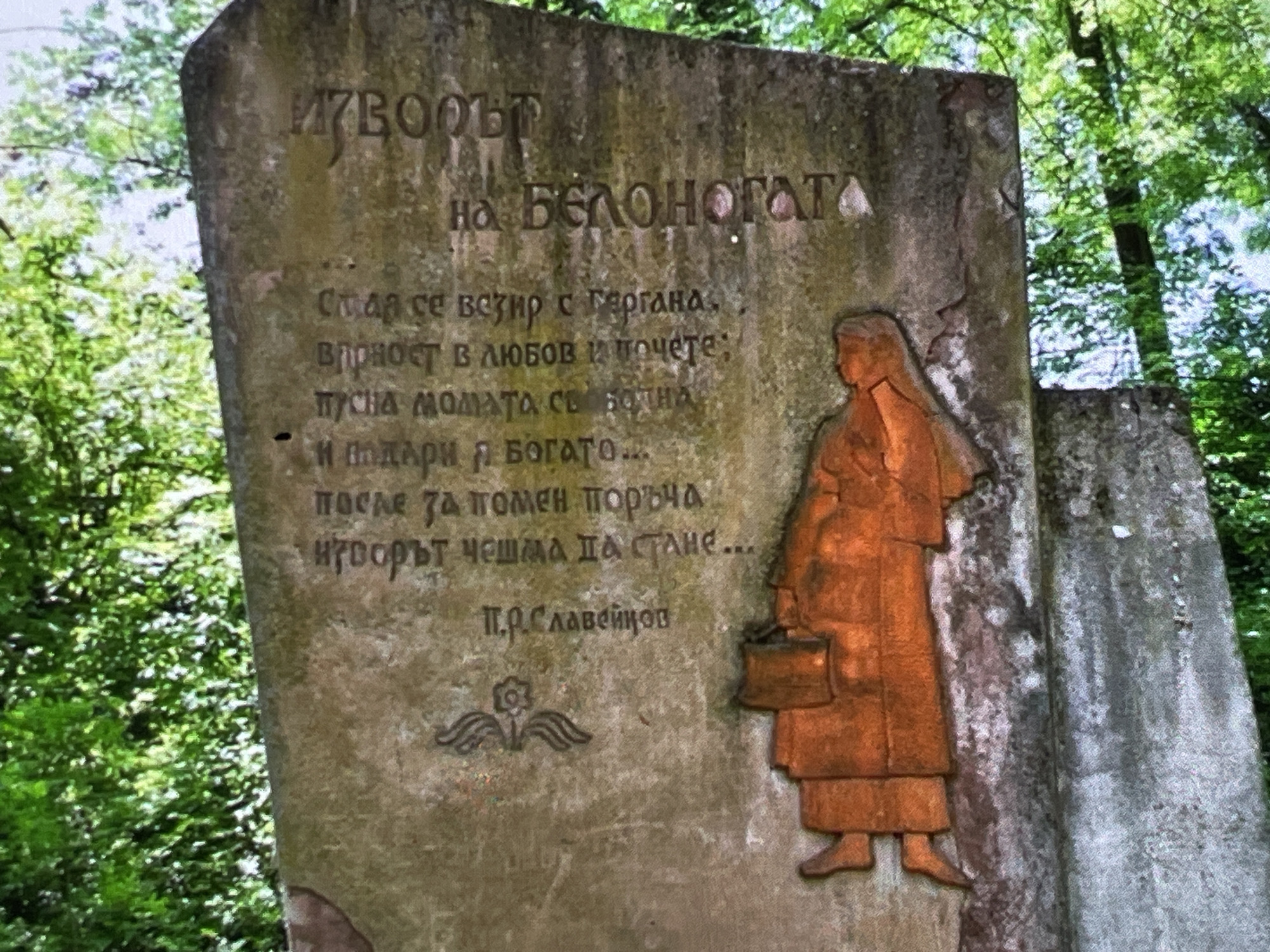 This is a close-up, slightly angled, eye-level outdoor shot of a weathered, stone monument in Harmanli, Bulgaria. The monument is carved with text in an unfamiliar alphabet and features a relief of a person dressed in an orange, robe-like garment. The person is depicted from the torso down, with legs and feet visible, and the figure is facing left. Above the figure and to the left, a small, stylized winged emblem is also carved into the stone. The text on the monument is inscribed in a Cyrillic-like script. The monument is set against a backdrop of lush green foliage from trees overhead, suggesting it is located in a natural, outdoor setting. The stone itself appears aged, with visible cracks and wear.