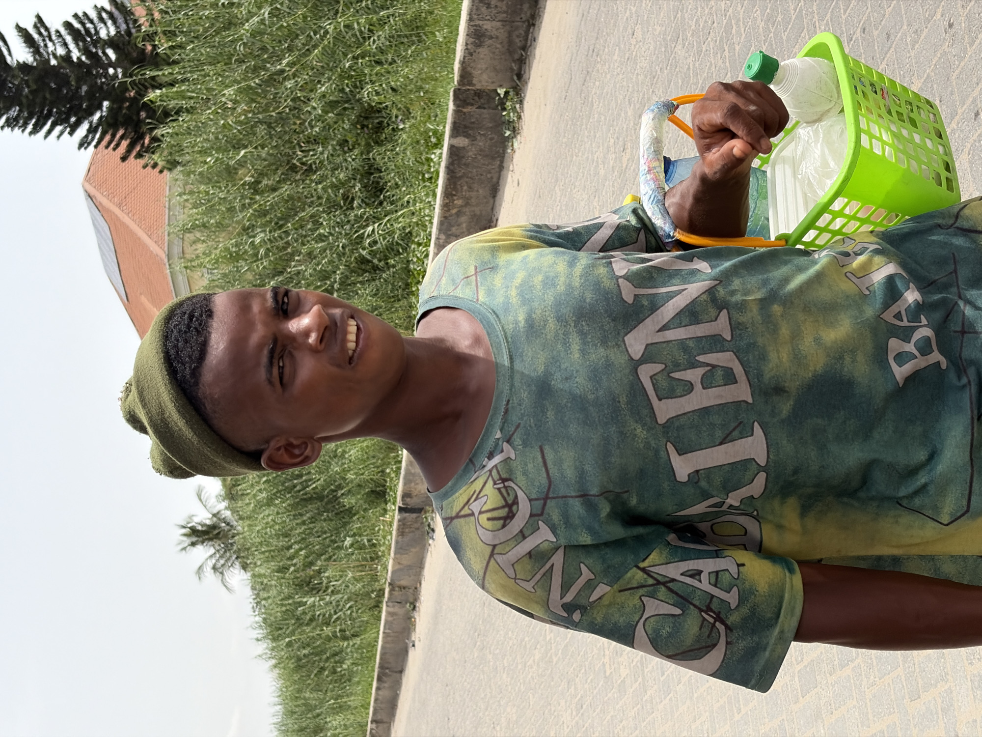 A young man stands outdoors on a paved surface, possibly a road or sidewalk, in Lekki, Nigeria. He is holding a green plastic basket filled with what appears to be bottled water and other items. The man is wearing a green t-shirt with large white lettering and a matching green beanie. The background includes lush green vegetation, a building with a red roof and solar panels, and a concrete curb. The weather appears sunny and clear.