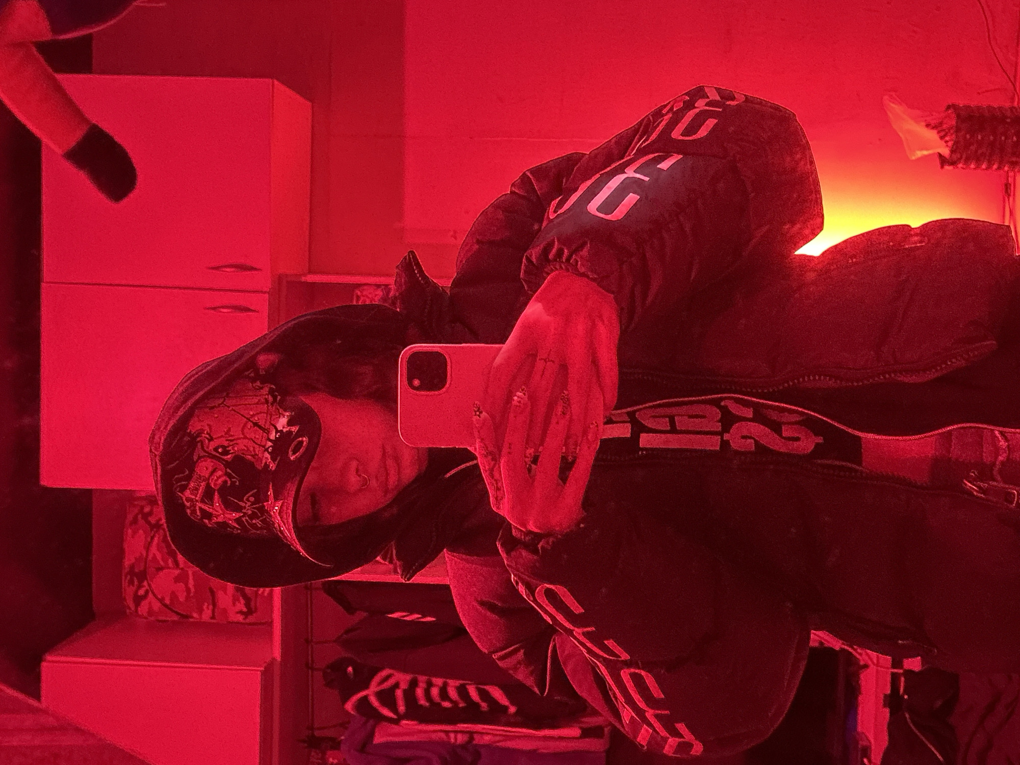The image is a vertical mirror selfie, rotated 90 degrees clockwise from its natural orientation, taken in an indoor setting bathed predominantly in intense red light, with a strong yellow-orange glow emanating from the right background. While the location context is Buenos Aires, Argentina, no specific geographical cues are visible in the image itself.
The subject is a person whose face is largely obscured by a light-colored smartphone they are holding to take the picture. They are wearing a dark, possibly black, puffy jacket or hoodie. Visible on the jacket are large, light-colored letters such as "JG" or "JC" on the upper chest area, and similar lettering further down. The hoodie also features an intricate, dark design, possibly a skull or abstract pattern, on its front. Around the person's waist or hip area, a dark band with visible text, including the number "2" and other indistinct letters, is apparent, suggesting it might be part of their clothing or an elastic band. The person's visible hand, holding the smartphone with a dual-camera system, shows what appears to be detailed nail art or rings.
In the background, to the left, a light-colored, possibly white, appliance resembling a small refrigerator or a two-door cabinet is prominent, with a smaller cabinet or shelf unit below it. On the far left, a dark, plush object, possibly part of a stuffed animal, can be seen. On the far right, near the intense yellow-orange light, there is a textured, possibly woven object.
The primary activity depicted is the taking of a mirror selfie. The time of day is not discernible due to the artificial lighting. The pervasive red light creates a moody and high-contrast atmosphere, strikingly contrasting with the warm, bright yellow-orange light from the background. No specific emotions are discernible from the partially hidden face.