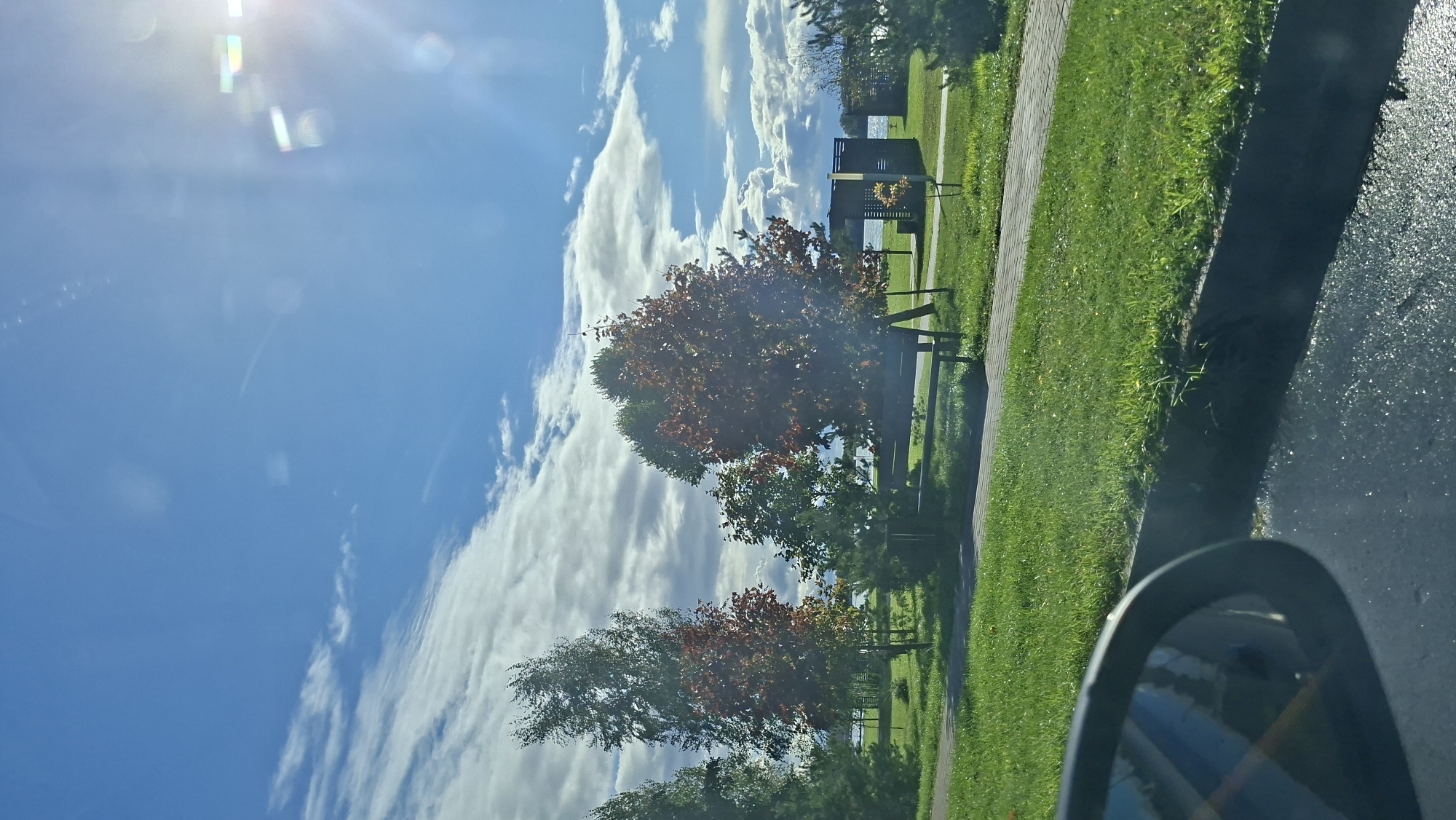 The image captures a sunny day with a blue sky filled with fluffy white clouds. The sun is bright and partially obscured by lens flare. In the foreground, a car's side mirror is visible on the left, reflecting a portion of the scene. A paved walkway runs through a grassy area with several trees. Some trees have started to show autumn colors with red and orange leaves, while others are still green. A dark-colored bench is placed on the grass next to the walkway. In the background, there's a glimpse of a body of water, likely a lake, and a small wooden structure, possibly a shelter or gazebo. The overall environment suggests a park or recreational area. The scene is consistent with a location in Myadzyel, Belarus, known for its natural beauty and proximity to lakes. No people are visible in the image.