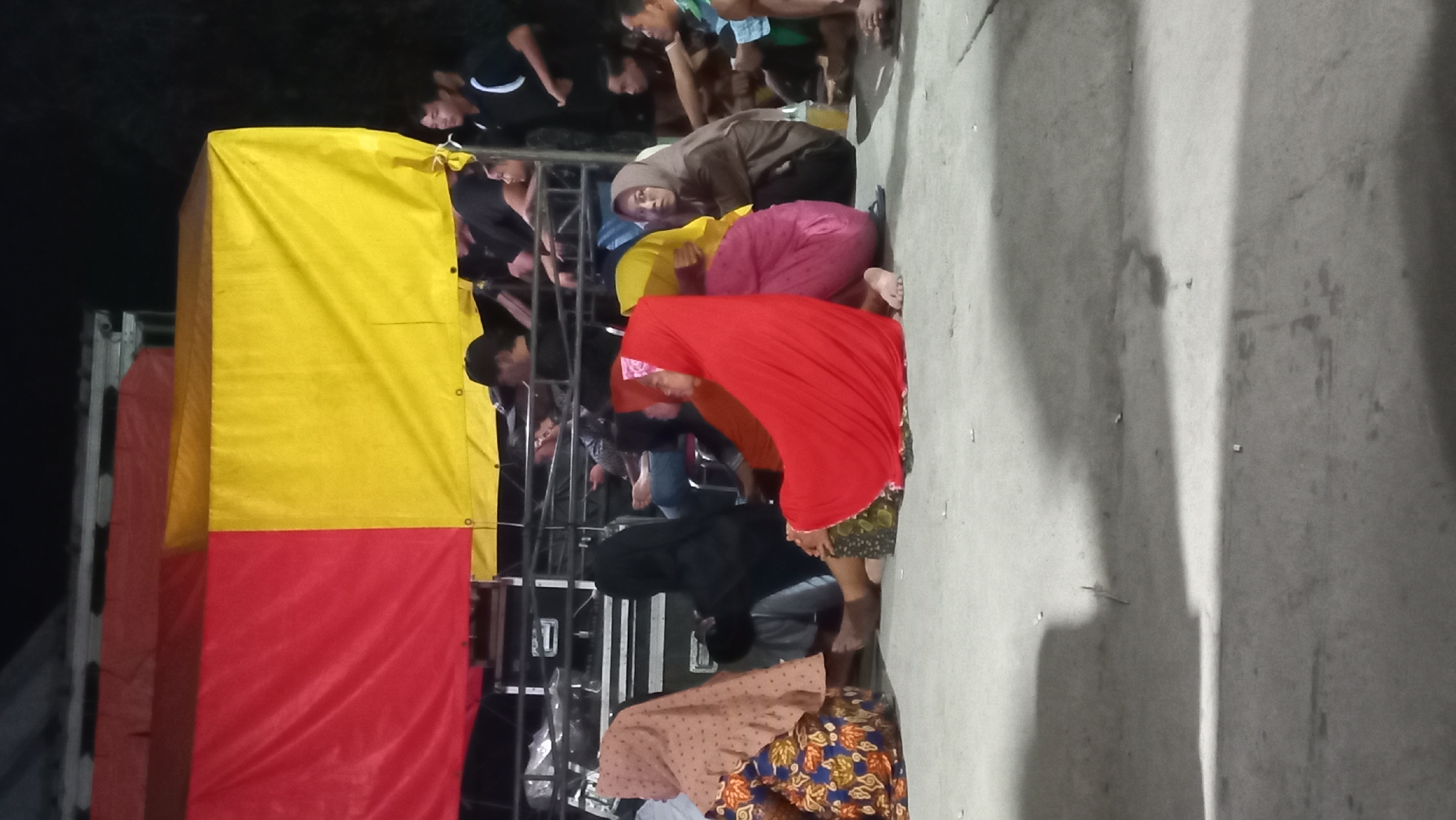 A nighttime outdoor scene shows a group of people seated on a concrete ground, some with their backs to the camera. Behind them, a stage setup is visible, draped with red and yellow tarpaulins. Several individuals are standing behind a metal barrier in front of the stage. The overall lighting is dim, suggesting it's night, and the ground appears to be a paved area, possibly a parking lot or an open space. The scene could be at a public event or gathering in Kedungmegarih, Indonesia.