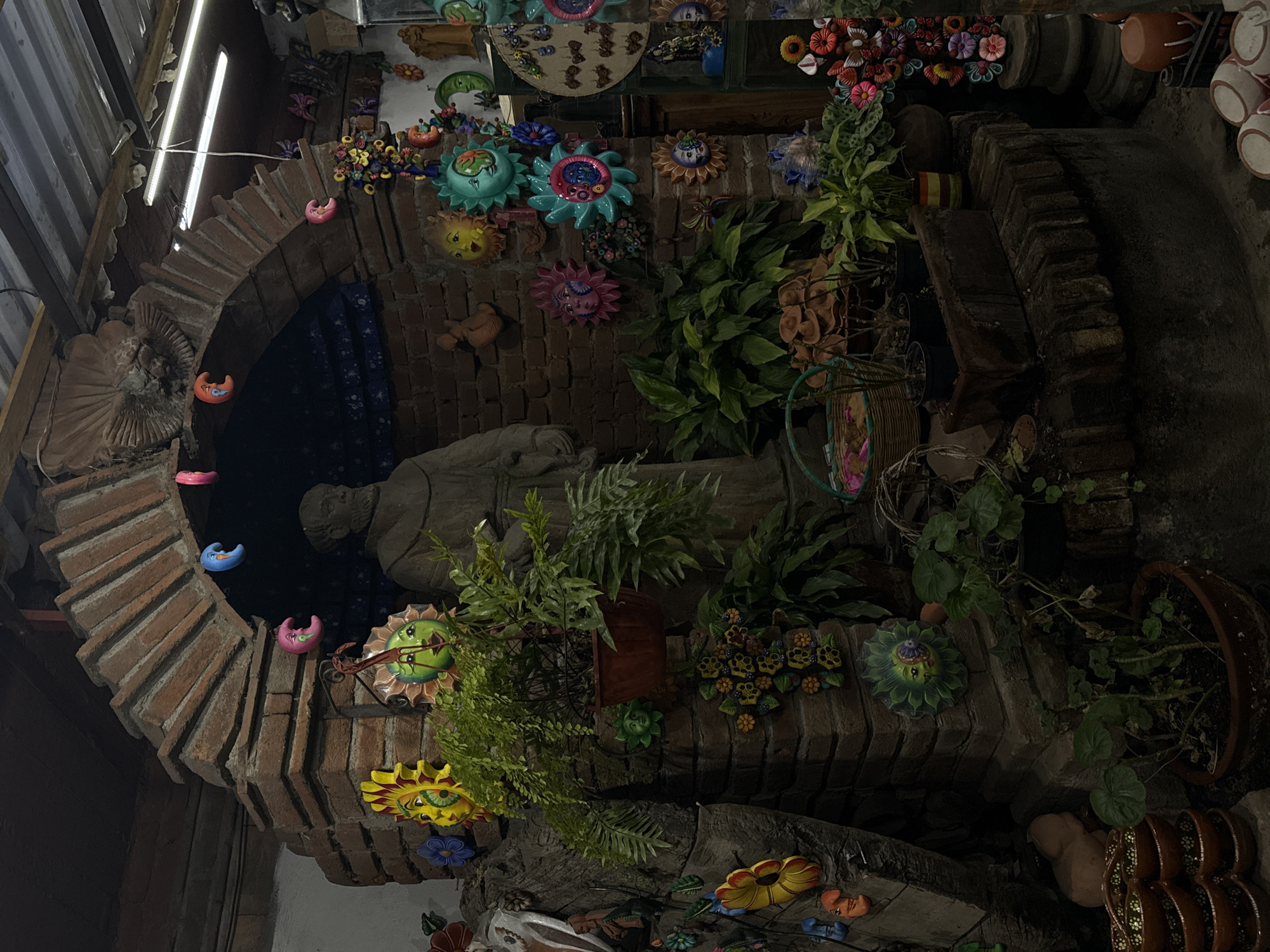 The image depicts an indoor display, likely a shop interior, showcasing handmade Mexican crafts in Metepec, Mexico. A brick archway frames a dimly lit alcove containing a light grey statue. The archway is adorned with colorful ceramic sun and moon decorations. The walls and structures around the arch are covered with various ceramic decorations, including flowers and faces, interspersed with potted plants and trailing vines. The overall impression is of a vibrant, crowded space filled with handcrafted items. A rack and lighting are visible in the upper left corner. The scene appears to be still life arrangement with no people or direct interactions depicted.