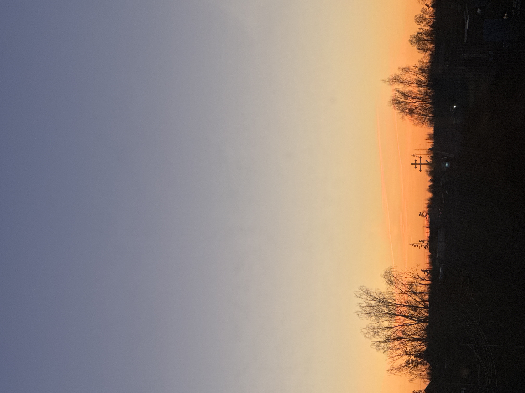 The image shows a sky at sunset. The left side of the image is a pale blue, transitioning to a yellowish-orange on the right. In the foreground on the right side are silhouettes of trees and possibly structures against the orange sky. Power lines are visible in the middle. The photo appears to have been taken outdoors, possibly in the outskirts of Фоки, Russia.