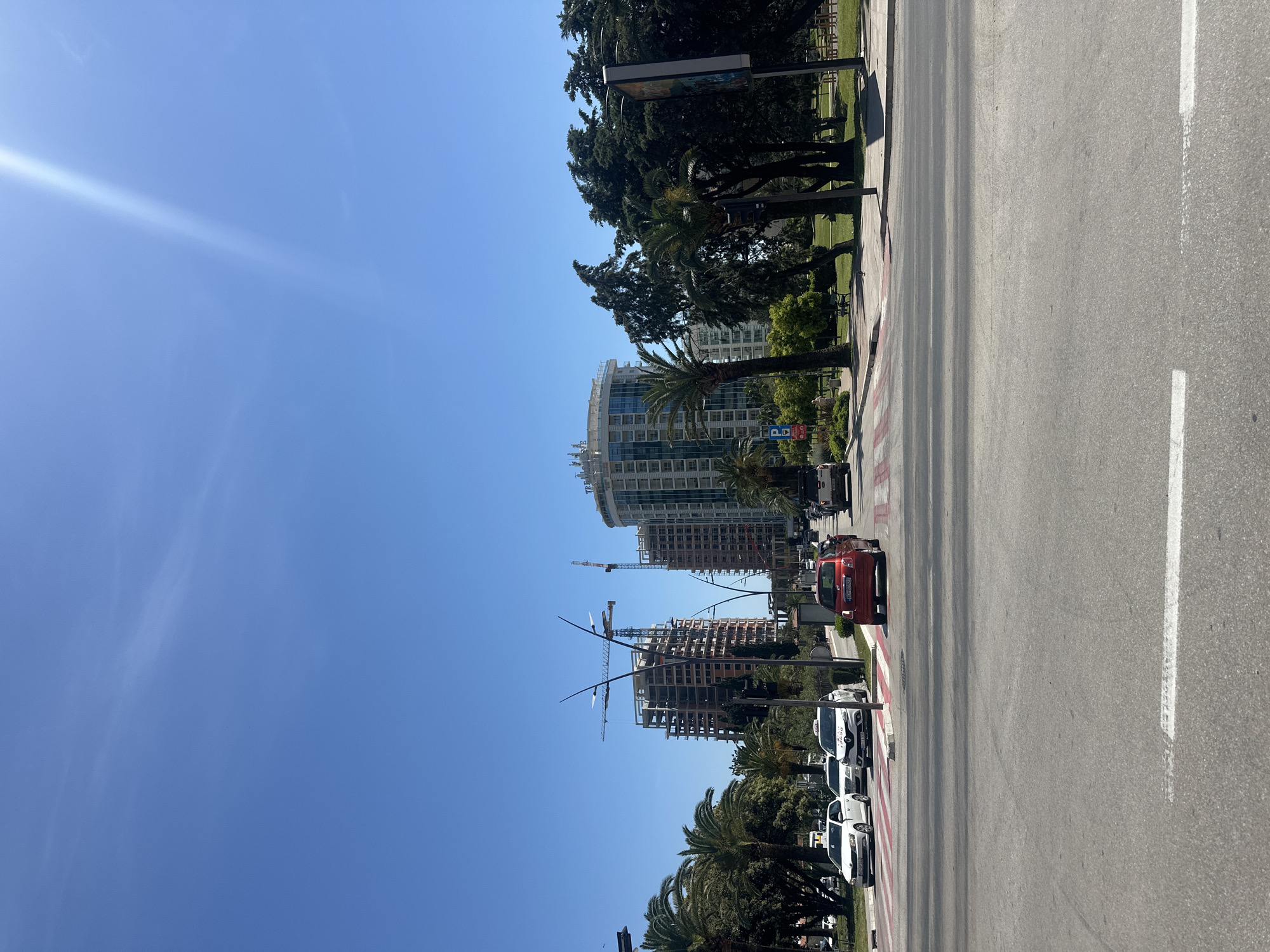 The image shows a street scene in Budva, Montenegro, taken on a sunny day. The view is oriented vertically, with the sky on the left and the road on the right. The sky is clear blue with a bright spot suggesting the sun's location. Along the left side of the road are trees, including palm trees, and several buildings. A building under construction is visible, with a crane adjacent to it. Completed high-rise buildings are further down the street. A row of parked cars lines the street. A red car is driving along the road, crossing a zebra crossing. The road itself is asphalt with painted white lines. A pedestrian crossing is marked by red and white stripes. The image captures a typical urban scene in Budva.