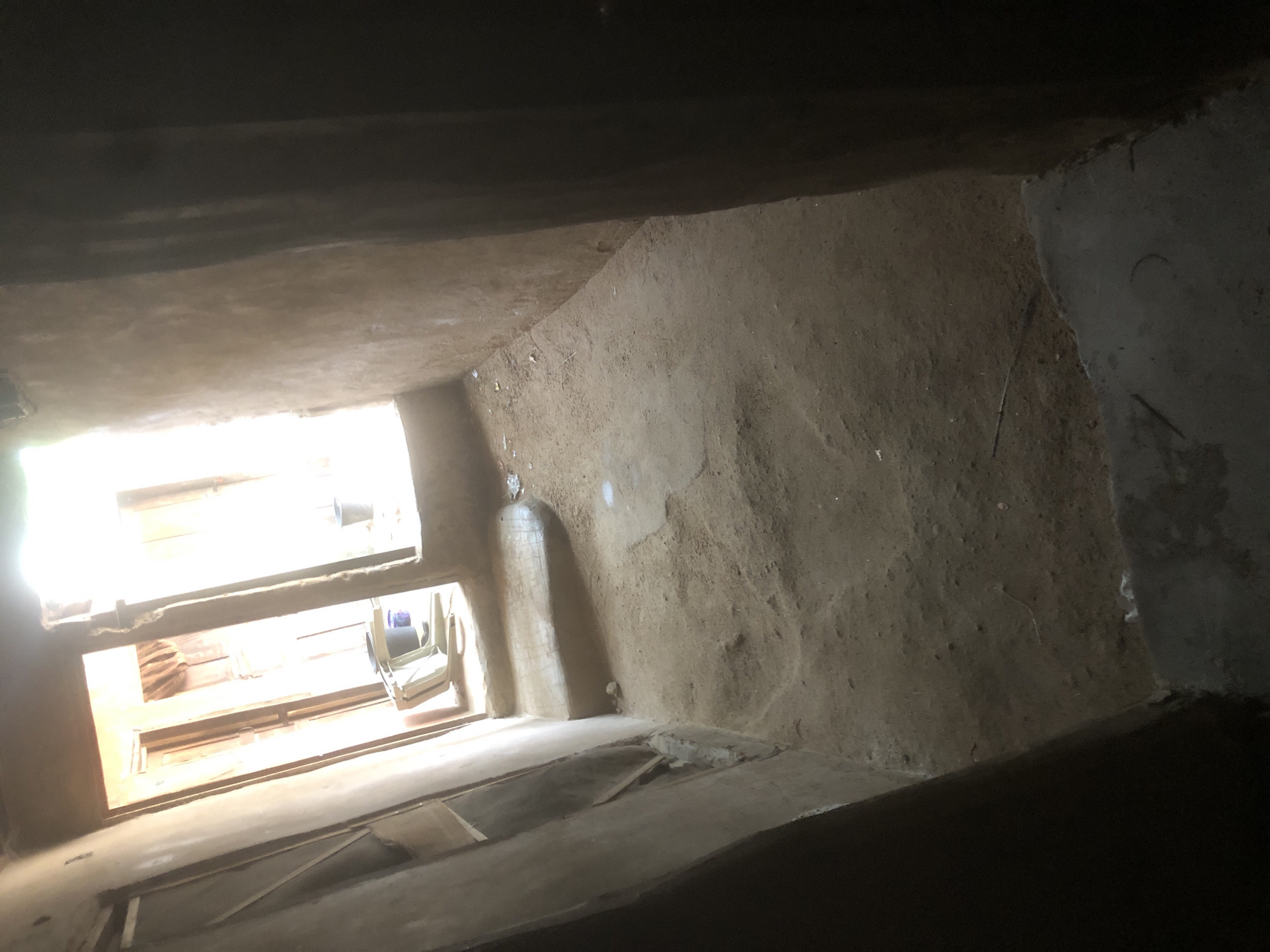 The image depicts an indoor corridor with a dirt floor and rough, unfinished walls. In the background, there are two doorways leading to brighter areas, possibly outdoors. Through one doorway, a plastic chair and a bucket are visible. The overall impression is one of simplicity and possibly poverty. The location is identified as a residential area in Jalingo, Nigeria.