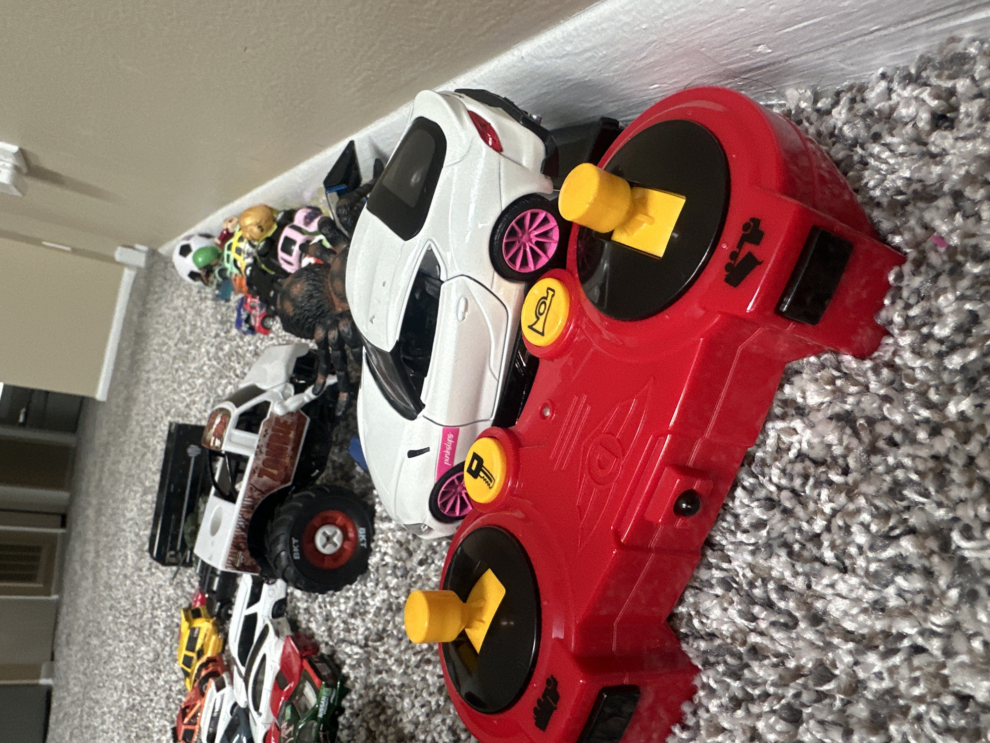 The image depicts a collection of toy vehicles and a remote control on a carpeted floor. In the foreground, a red remote control with black accents and yellow joystick tops is visible. Behind it, a white toy car with pink rims and a "pinkslips" decal is positioned. Further back, a white monster truck toy with "ZOMBIE" emblazoned on its side and large black tires with red rims is present. Scattered around are other toy cars of various colors, including yellow, red, and green, as well as a toy soccer ball. The environment appears to be an indoor setting, likely a child's play area, with a carpeted floor and a baseboard visible on the right side. The lighting is neutral. The image does not provide information about the city of Schaumburg, United States of America, or any specific location within it.