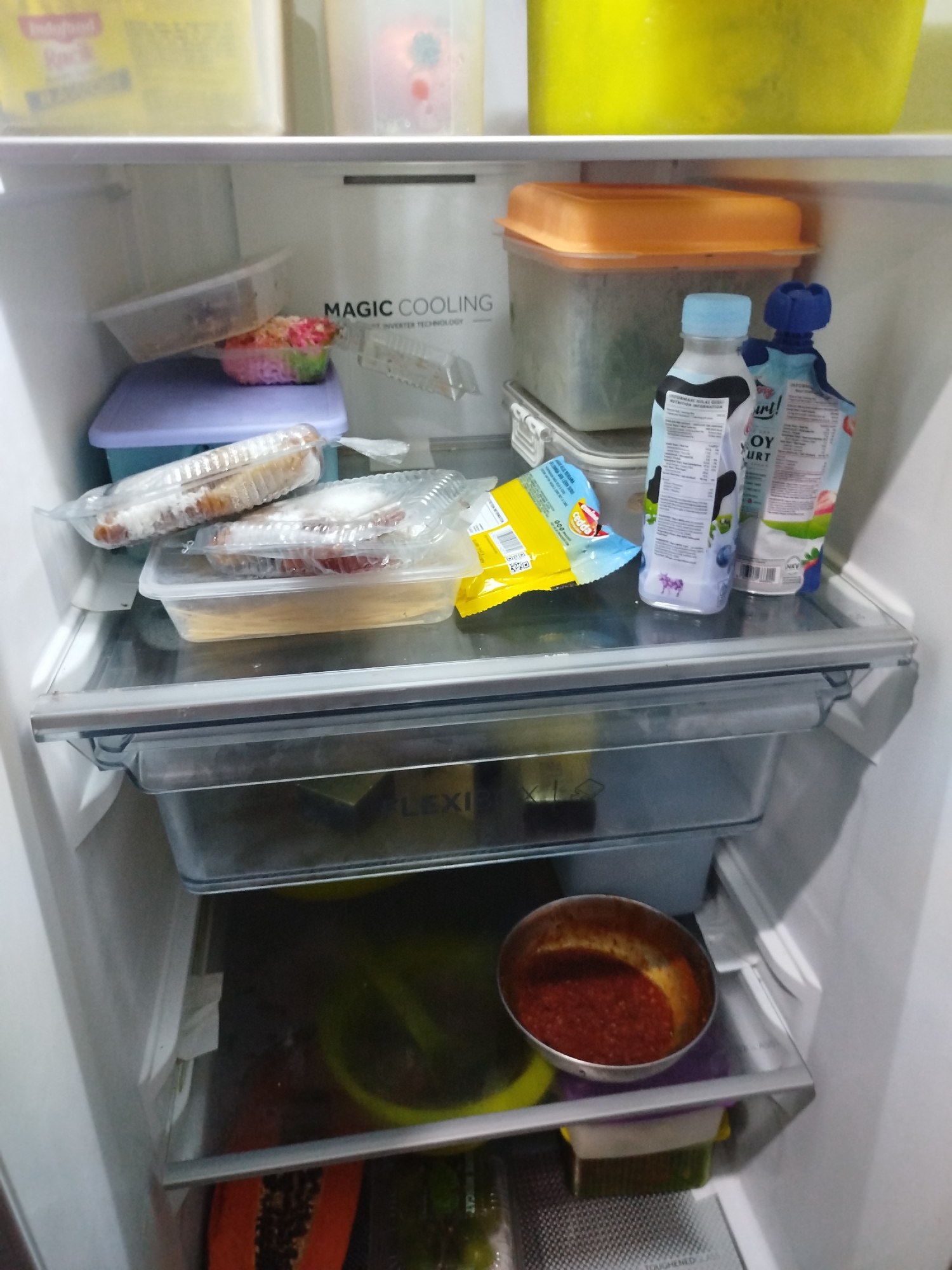 The image depicts the interior of a refrigerator, likely located in a residential kitchen in Bandungrejo, Indonesia. The refrigerator contains various food items and containers organized on its shelves. On the top shelf, there are two large yellow and green containers. Below, on the second shelf, there is a clear plastic container with a pink and colorful salad or dessert, a clear plastic container with what appears to be food wrapped in plastic, and several other plastic containers, some with lids. Among these, there are two bottles of yogurt, one blue and one with a blue cap, and a yellow packet of snacks. The third shelf holds two more plastic containers of food, one with spaghetti and another with some type of pastries or buns. The bottom shelf contains a metallic bowl filled with a red sauce or paste, a green bowl, and a piece of papaya with its seeds visible. Additional containers and items are visible in the drawers at the bottom of the refrigerator. The environment is well-lit, suggesting the refrigerator is in use.