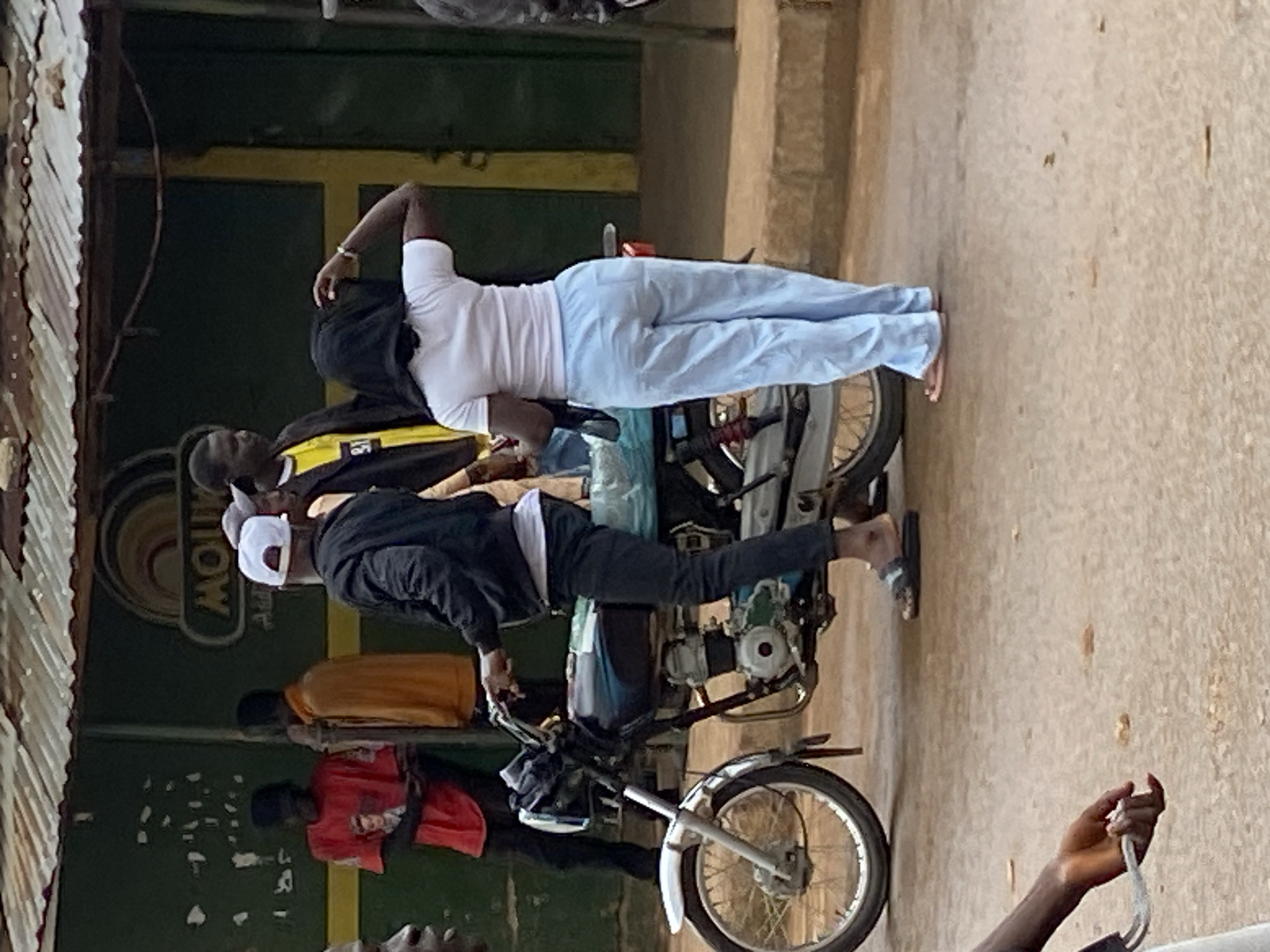 The image depicts a street scene in Bukuru, Nigeria, with several people and a motorcycle. A man in a black jacket and a white cap is seated on a motorcycle, holding the handlebars. Next to him, a woman in a white t-shirt and light blue pants stands with her back to the camera, touching her head. In the background, two men are visible; one is wearing a red t-shirt, and the other is in an orange jacket. A building with a corrugated metal roof and a green facade stands behind them. The ground is paved with concrete and dirt.