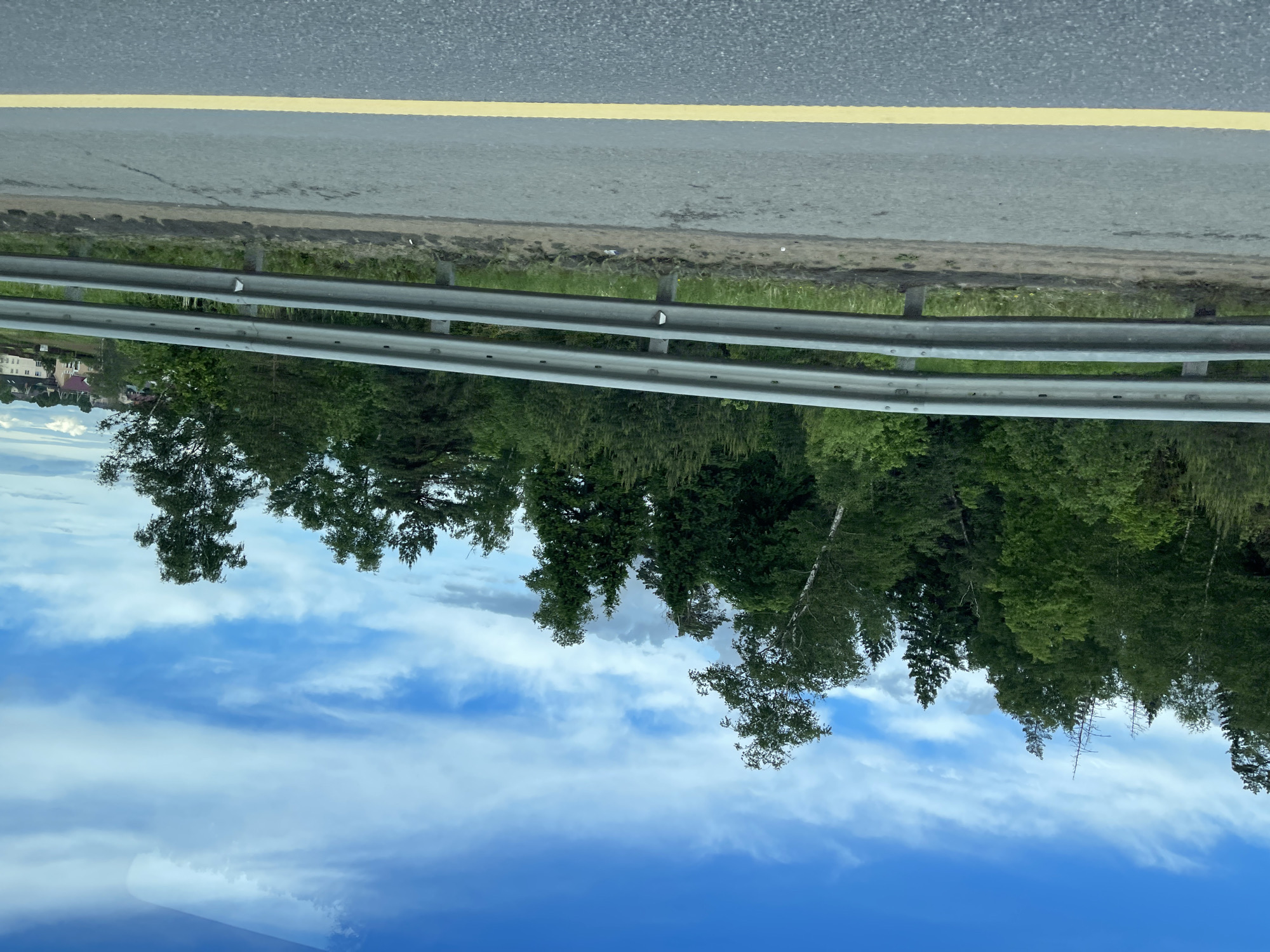The image shows a view from a moving vehicle on a road in Бавыкино, Russia. The road is marked with a yellow line, and a metal guardrail separates the road from a forest. The sky is blue with scattered clouds. Some buildings can be seen in the distance.