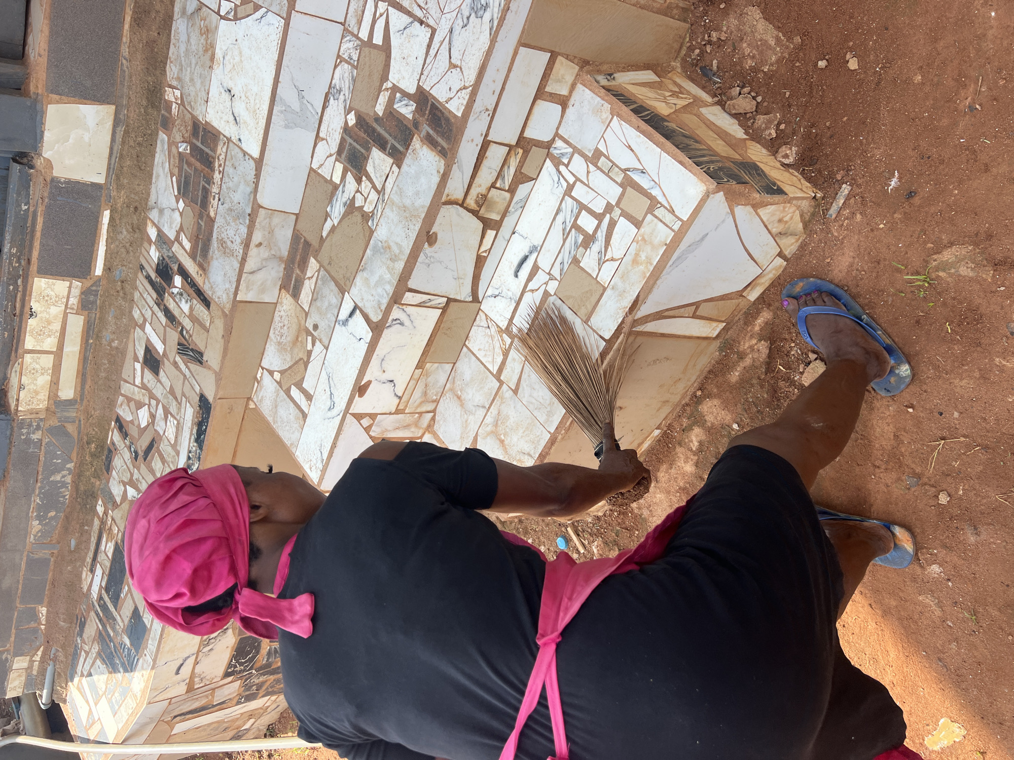 A person is sweeping a mosaic tiled outdoor staircase. The person is wearing a black t-shirt, a pink headscarf, a pink apron, and blue flip-flops. The staircase is constructed from various sizes and shapes of mosaic tiles, including white, beige, black, and marbled tiles. The ground is composed of reddish-brown soil with scattered small rocks and dry vegetation. The location is identified as Nkpor, Nigeria, suggesting an outdoor setting in a community or residential area. The lighting suggests it is daytime.