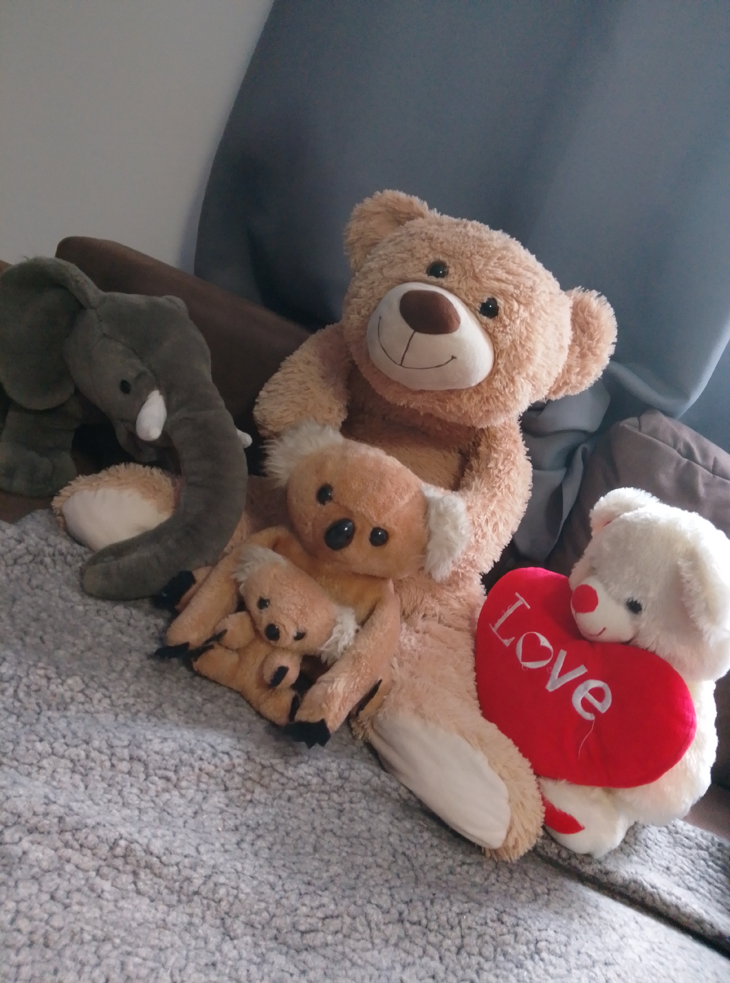 The image shows a collection of stuffed animals on a couch in an indoor setting. The stuffed animals include a large tan teddy bear, a grey elephant, two koala bears, and a white bear holding a red heart with the word "Love" embroidered on it. The couch appears to be covered in a textured grey blanket. The background consists of a blurred curtain and cushions. The location is identified as being in the city of Wedel, Germany. There are no people or actions depicted in the image, only a static arrangement of stuffed toys.
