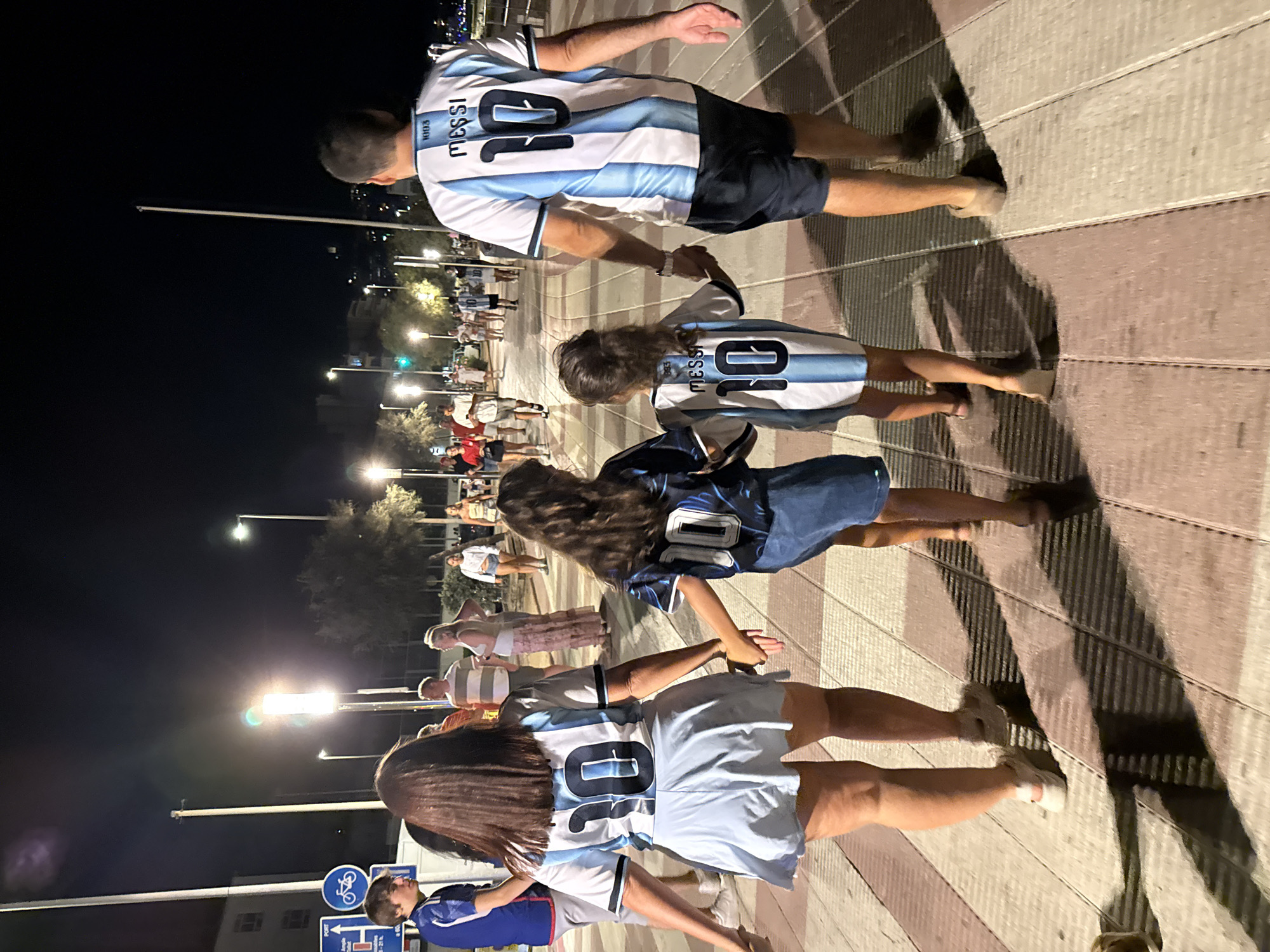 In Blanes, Spain, at night, a family walks along a pedestrian promenade. The promenade has wooden planks and is illuminated by streetlights. A man and a child, possibly father and daughter, wear matching light blue and white striped jerseys with "MESSI 10" printed on the back. The man is wearing dark shorts, and the child is wearing a denim skirt. Beside them, a woman, presumably the mother, also wears a similar jersey with "10" visible, paired with a light blue skirt. Another child, in a dark jersey with "10" on the back, walks between the woman and the man. Other people are visible in the background, some appearing to be tourists. There is a blue sign with a bicycle symbol and text in Spanish on a pole.
