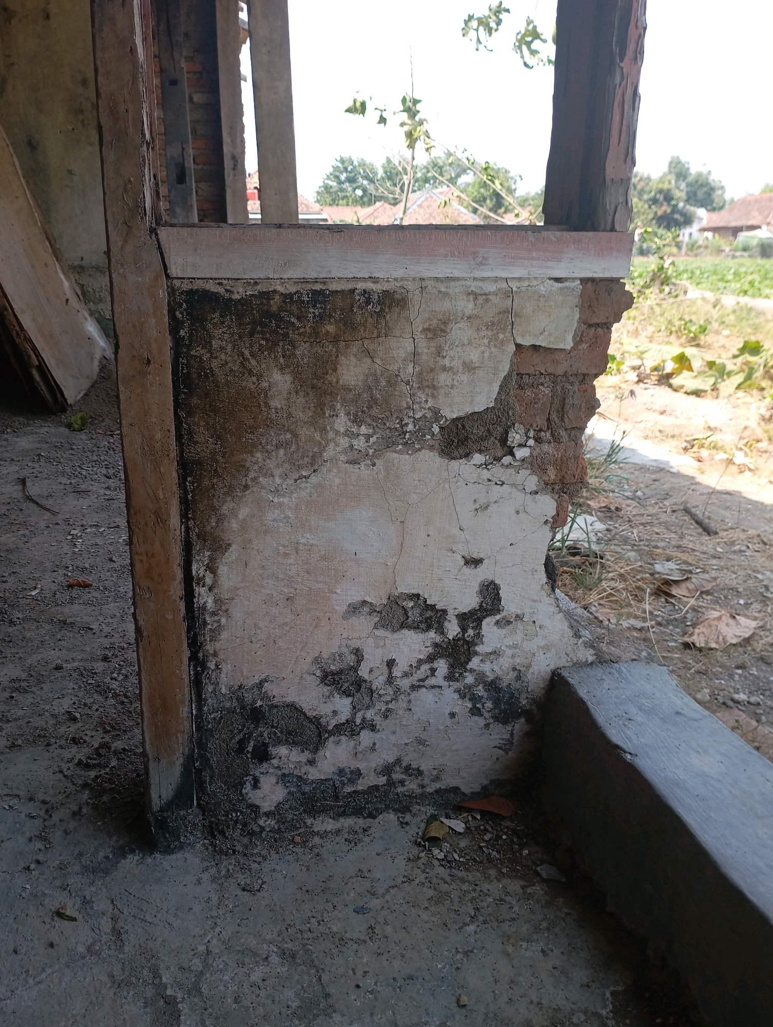 The image depicts a weathered, crumbling section of a building's exterior in Kecamatan Haurgeulis, Indonesia. A wooden post stands vertically on the left, adjacent to a wall made of brick and plaster. The plaster is heavily cracked and stained, with large sections missing, exposing the underlying brickwork. The ground surrounding the structure is rough concrete and gravel, with scattered dry leaves and debris. In the background, a glimpse of a rural landscape is visible, with trees and houses under a bright, hazy sky. The scene suggests neglect and disrepair.