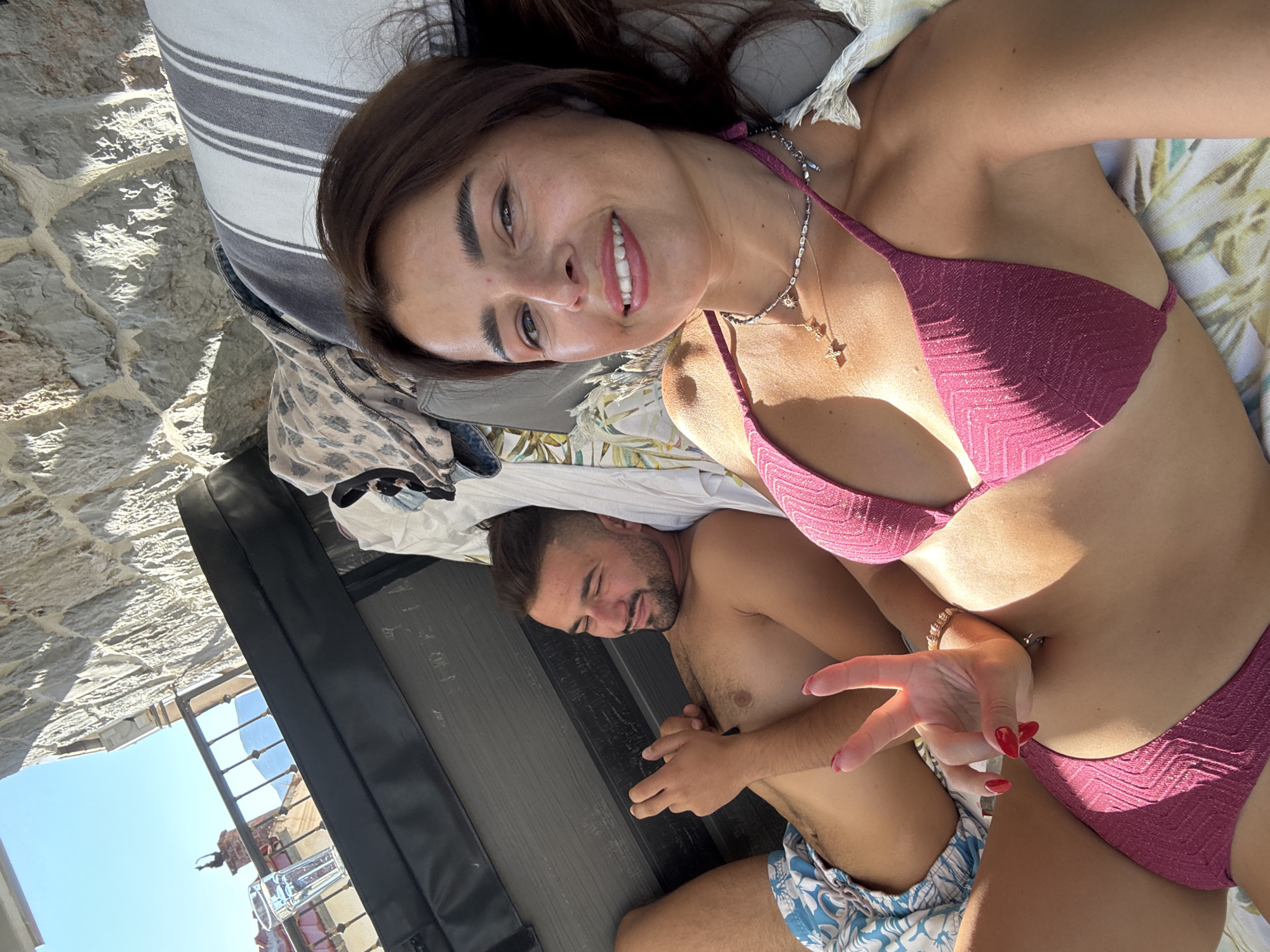 A woman in a pink bikini smiles at the camera while holding up a peace sign with her right hand. She is lying down with a man in blue patterned swim trunks next to her. He is looking at his phone. They are on a lounge chair or sofa, with a stone wall and balcony visible in the background. Sunlight streams in from the right, casting shadows on the stone wall. A glass of water is on the balcony railing.