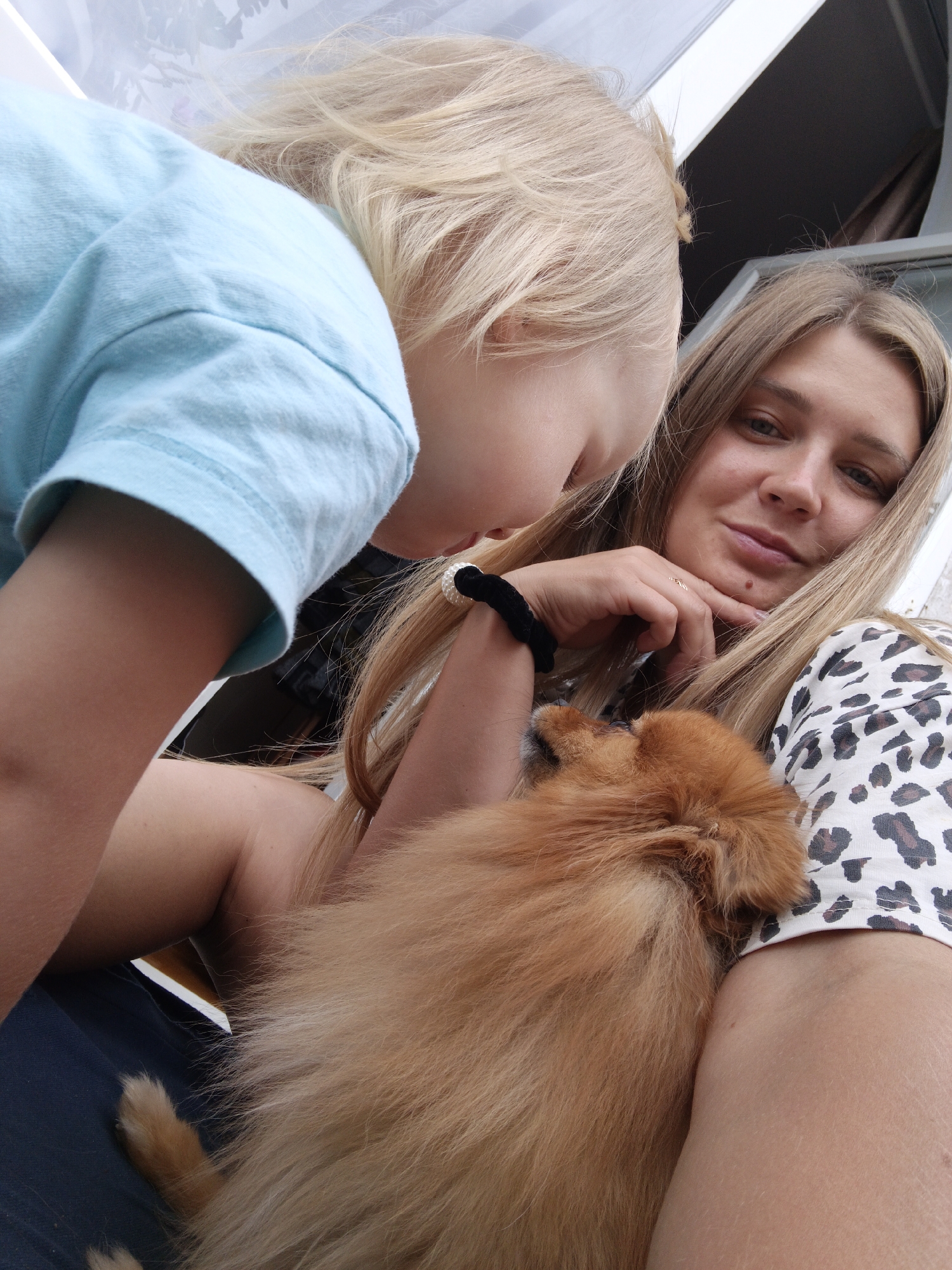 In this selfie taken outdoors, likely in Medvedyovka, Russia, a young child with blonde hair, wearing a light blue t-shirt, leans towards the camera, their face obscured. The child's arm, visible from the elbow down, is lightly touching a small, fluffy, light brown Pomeranian dog being held by an adult. The adult, a woman with long blonde hair, looks directly at the camera with a slight smile. She is wearing a white top with a black and brown leopard print pattern. Her right hand is resting on her chin, and she is wearing a thin gold ring on her ring finger. The dog is positioned in the crook of her arm. The background is blurred, but hints of outdoor scenery and possibly a canopy or awning are visible. The overall impression is a candid moment of affection between the child, the adult, and their pet.