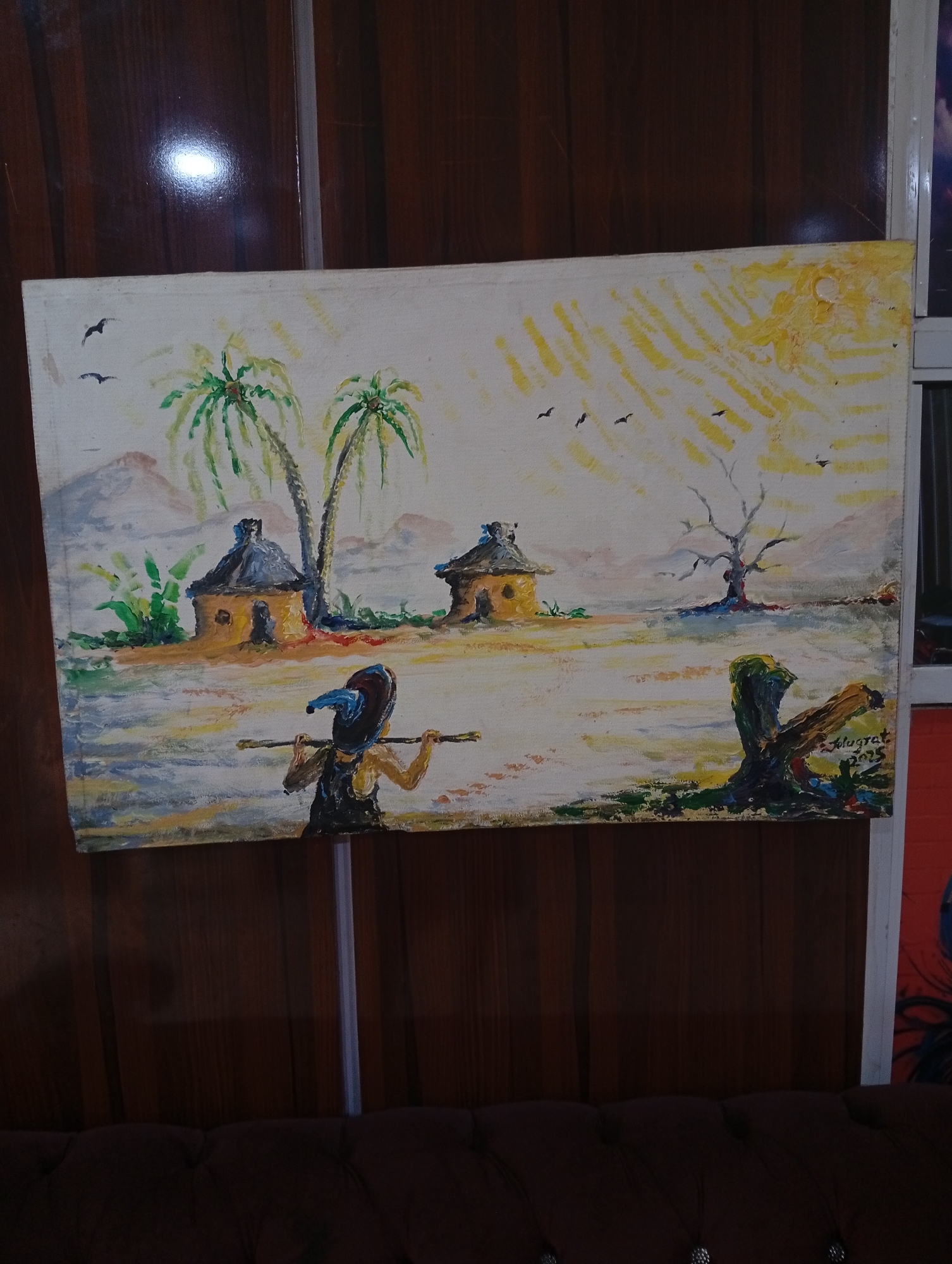 The image shows a painting depicting a rural scene in Adankolo, Nigeria. The painting includes:
* Two round huts with conical roofs.
* Two palm trees.
* A bare tree with several birds flying overhead.
* A person in the foreground carrying something (possibly a yoke) on their shoulders.
* An object that appears to be a tree stump in the foreground to the right.
* The painting is dated "2015".
* The background shows hills.
* Above the hills is a hazy or sunny sky with indications of a sun.
The painting is mounted on a wall with vertical wooden paneling. A brown leather sofa is in the foreground.