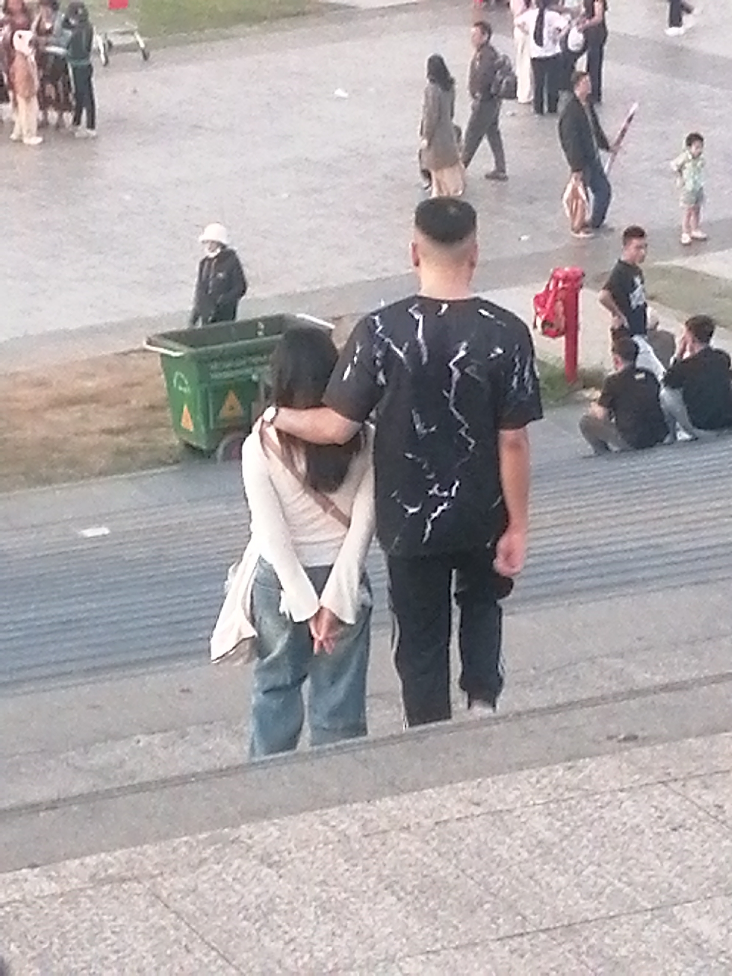 The image depicts an outdoor scene in a city, possibly Da Lat, Vietnam, given the architectural style and general ambiance. In the foreground, a couple is walking up a wide set of stone steps. The male is in front, wearing a black t-shirt with a white abstract pattern and black pants. The female, behind him, has her arm around his shoulder and is wearing a light-colored long-sleeved top and blue jeans. Her hands are clasped behind her back.
Further up the steps and to the left, a person in a dark jacket and white helmet is standing near a green trash bin. In the background, there are several other people walking and standing around. One person, seen from the back, is carrying a red object. Another individual is dressed in a patterned shirt and shorts. The ground surface appears to be paved, with some areas of greenery visible. The lighting suggests it is daytime, possibly late afternoon.