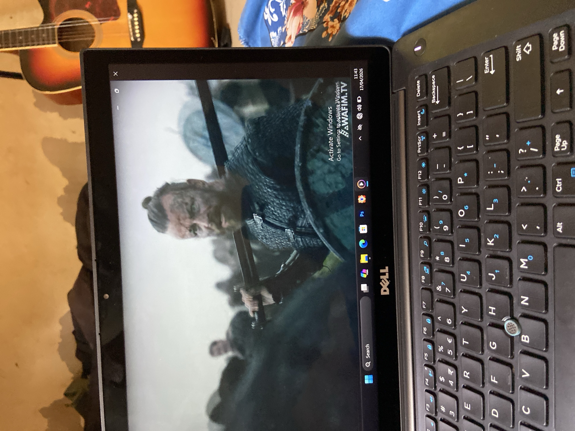 An upward-tilted shot captures a Dell laptop opened and displaying a video, with a keyboard visible to the right. The image is rotated 90 degrees clockwise.
On the laptop screen, a fair-skinned person, likely a man with a beard and intense expression, is shown from the chest up, holding a sword. He is dressed in what appears to be chainmail armor, suggesting a historical or fantasy setting. In the upper right corner of the screen, text reads "Activate Windows Go to Settings to activate Windows." Below this, "BAWAFIM.TV" is visible, along with the current time "11:43" and date "17/04/2026." The Windows taskbar is at the bottom of the screen, showing a search bar, Windows icon, and various application icons including Photoshop (Ps), a browser, and a file explorer. System icons for volume, Wi-Fi, and battery are also present next to the date.
The laptop's black keyboard features a QWERTY layout with numeric keys and function keys (F1-F12). Some keys display additional symbols like the Euro (€) and Indian Rupee (₹) symbols. A small, blue-grey joystick-like pointing device is integrated into the 'B' key. The Dell logo is visible on the laptop's bezel.
In the background, partially obscured, is an acoustic guitar with a sunburst finish, leaning against a light-colored surface. The overall scene suggests an indoor setting where someone is watching content on a laptop. The time and activation prompt indicate personal computer use. The location context for this media is Mararaba, Nigeria.
