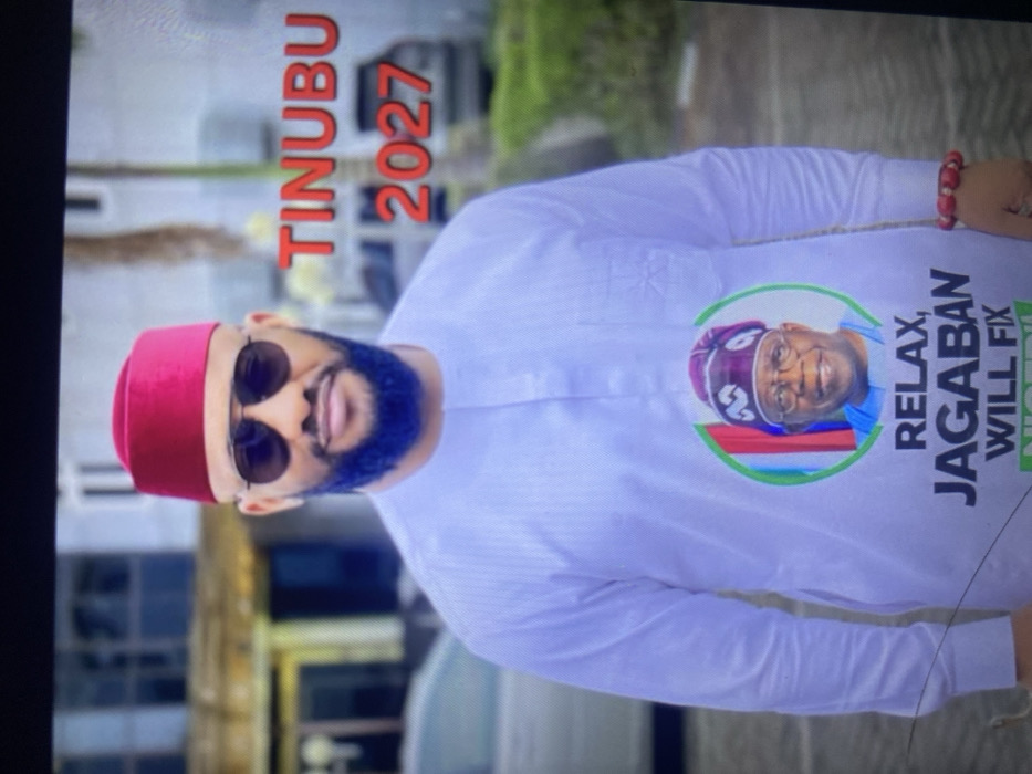 A man with a blue beard and sunglasses wears a red fez and a white shirt. The shirt features a button with a picture of a man wearing a purple cap, and the text "RELAX, JAGABAN WILL FIX NIGERIA". The text "TINUBU 2027" is visible in red above the man's head. He is wearing a beaded bracelet. The background is blurry, suggesting an outdoor setting.