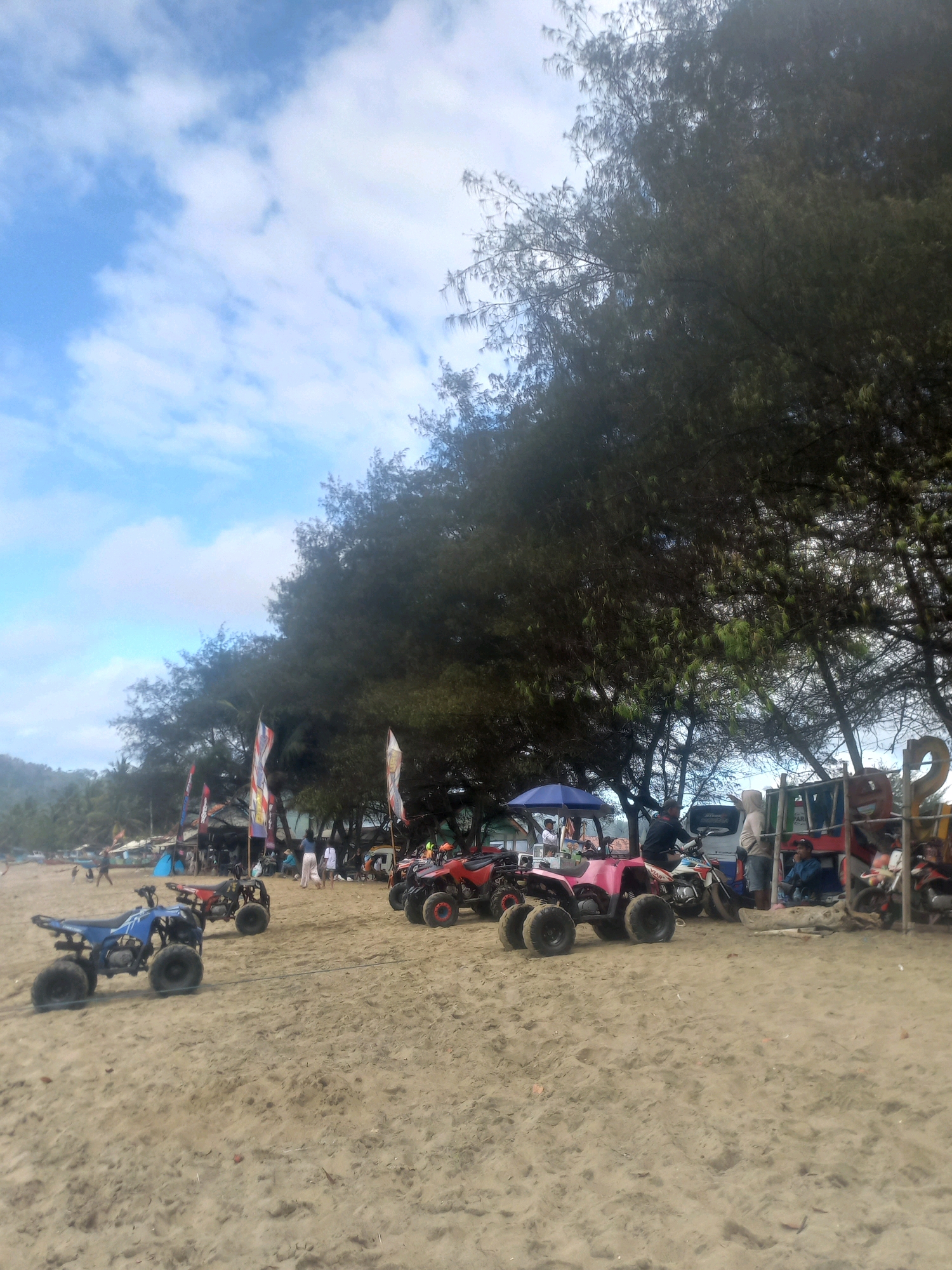 The image depicts a sandy beach in an unidentified location in Indonesia, likely a tourist area due to the presence of ATVs for rent and small food stalls. In the foreground, several ATVs are parked on the sand, including a blue ATV on the left and pink and red ATVs further back. Some people are visible in the background, including individuals standing near stalls and some riding a motorcycle. Tall trees with dense foliage line the back of the beach, providing shade. Colorful flags are also planted along the stalls. The sky is partly cloudy with patches of blue. The overall impression is of a relaxed, recreational setting by the sea.
