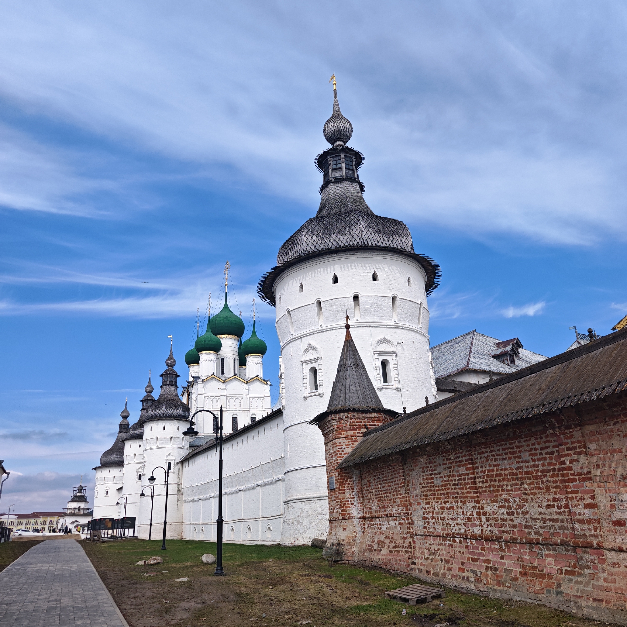 The image captures a portion of the Rostov Kremlin, a historic architectural complex located in the city of Rostov, Yaroslavl Oblast, Russia. The perspective is at ground level, showcasing a paved walkway to the left and a grassy area with scattered vegetation.
Dominating the scene is a long, whitewashed wall punctuated by towers with dark, onion-shaped domes. These domes are tiered, with the bottom tiers darker in color, and appear to be made of wood. The wall extends into the distance, with additional towers and green-domed churches visible. To the right of the whitewashed wall is a shorter brick wall, exhibiting a weathered appearance, likely indicating age.
Several black lampposts are situated along the walkway, adding a modern touch to the ancient setting. The sky is blue with streaky clouds, providing a backdrop of fair weather conditions. There are no people visible in the foreground.