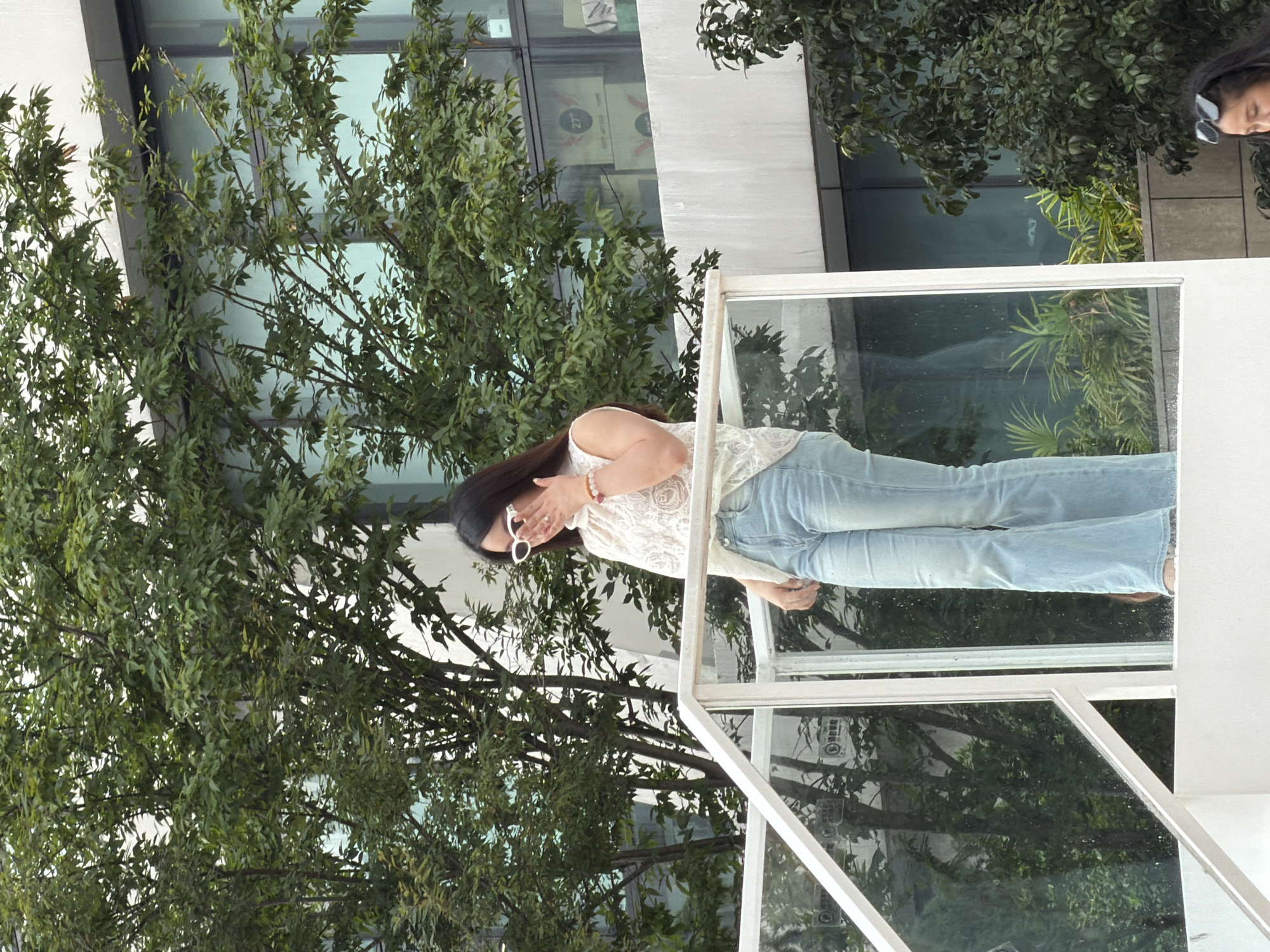 A woman stands on a white elevated platform with a glass railing. She is wearing a white lace top and light blue flared jeans, with white sunglasses pushed up on her head. Her right hand is raised to her face, touching her sunglasses. Behind her, green trees with lush foliage are visible, with a modern building featuring large windows in the background. Some boxes with logos are stacked on a ledge within the building's view. The environment appears to be an outdoor urban setting, potentially in Yuzhong District, China.