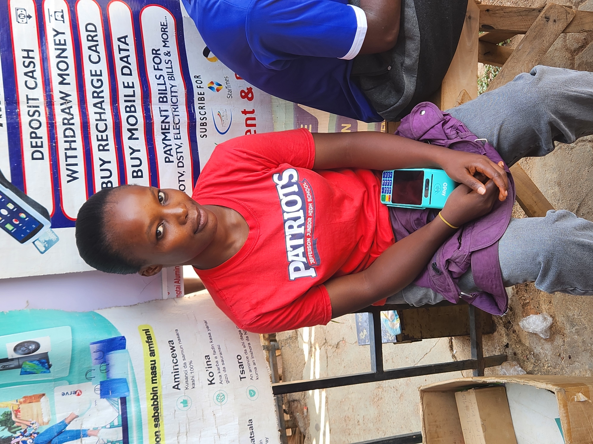 The image shows a young woman sitting outdoors in an unknown location. She is wearing a red T-shirt with "PATRIOTS JEFFERSON JUNIOR HIGH SCHOOL" printed on it and grey trousers. She is holding a light blue Opay POS device in her hands. Behind her is a sign advertising services such as "DEPOSIT CASH," "WITHDRAW MONEY," "BUY RECHARGE CARD," "BUY MOBILE DATA," and "PAYMENT BILLS FOR OTV, DSTV, ELECTRICITY BILLS & MORE...". Part of another sign promoting "AMINCEWA", "KO'INA" and "TSARO" is also visible. The setting appears to be a local business or kiosk. Another person wearing a blue shirt sits to her side, slightly out of focus. A cardboard box sits below the chair. The ground appears to be dirt with small pieces of trash.