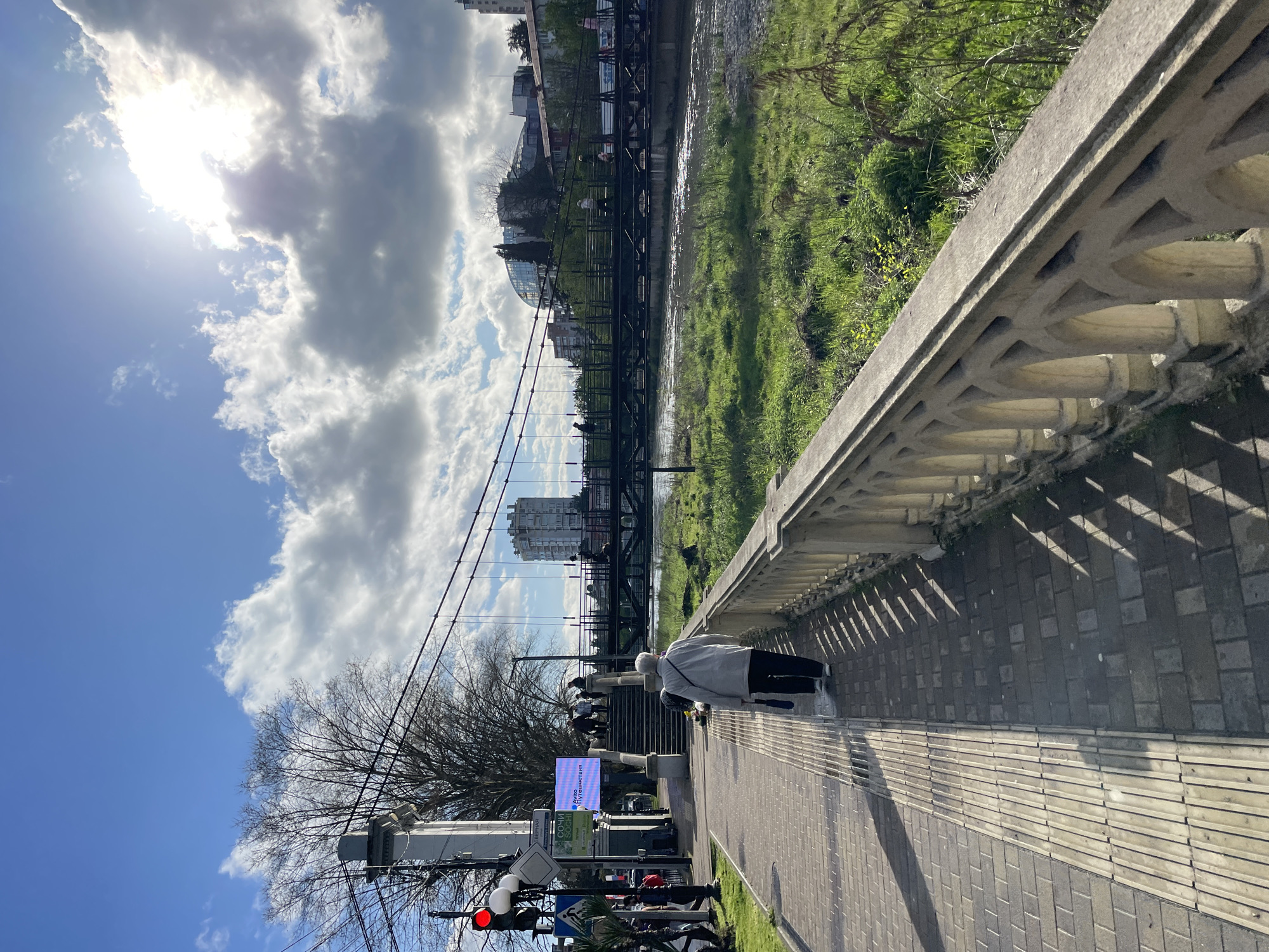 The scene is an outdoor shot in Sochi, Russia. A pedestrian walkway paved with rectangular blocks is lined by a low concrete balustrade with decorative openings. A woman in a light-colored coat and dark pants is walking along the sidewalk. To the left of the walkway are traffic signs, utility poles, and bare trees. A bridge spans a river that flows horizontally in the background. The sky is partly cloudy with the sun shining. A building with a glass dome roof is partially visible near the river. In the middle distance, there are people on the bridge, and taller buildings can be seen beyond. The image is rotated 90 degrees counterclockwise.