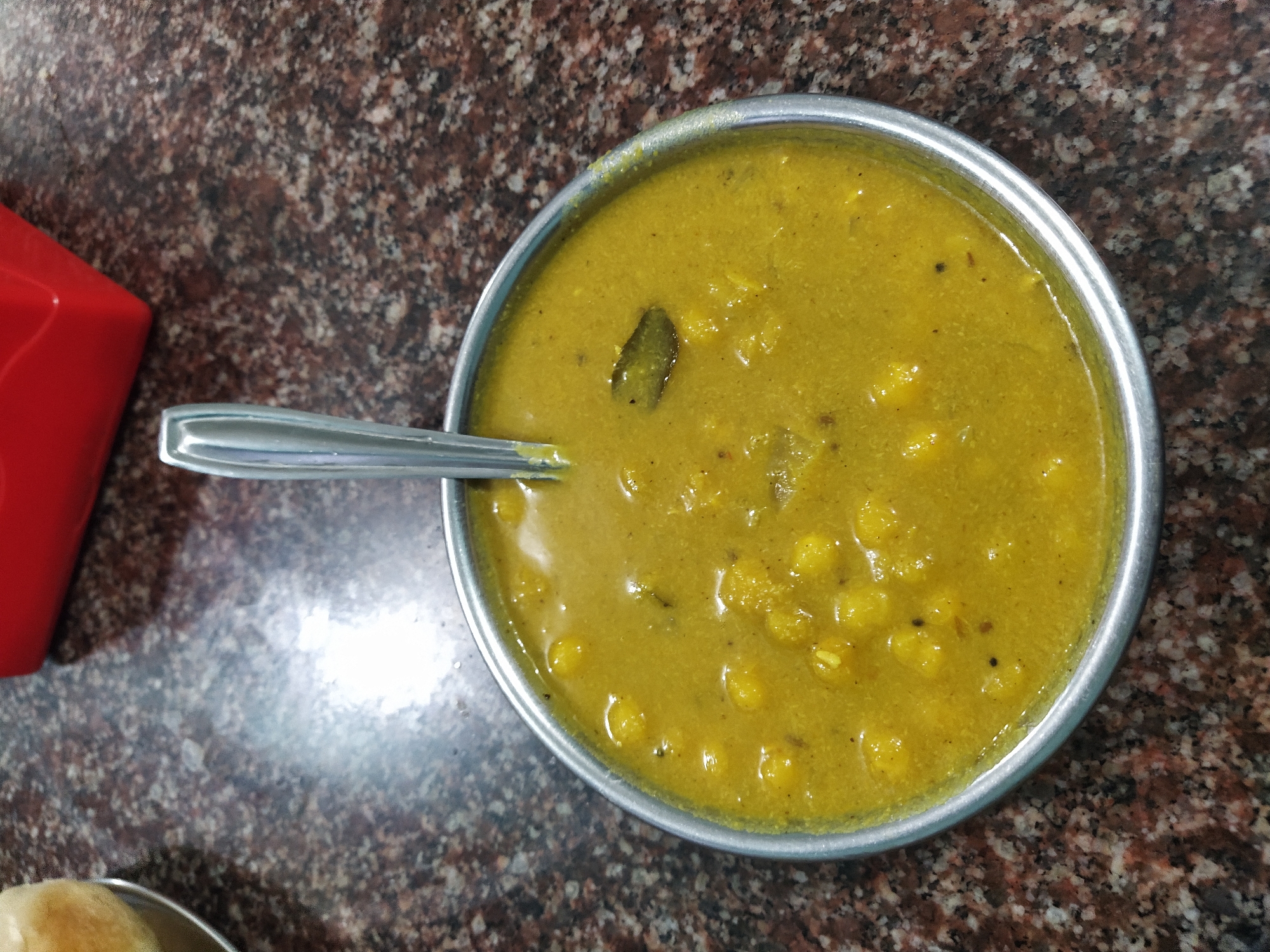 The image depicts a bowl of yellow chickpea curry (chole) with a spoon resting in it, placed on a dark speckled countertop. A red container is partially visible in the left corner. The bowl and spoon are made of stainless steel. A piece of dough can be seen at the bottom left corner, as well. The setting appears to be in a restaurant or eatery in Oxel, India. No people are visible.