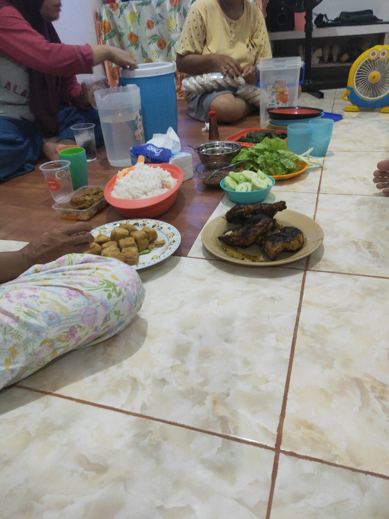 A group of people are seated on a tiled floor, sharing a meal. The environment appears to be indoors, likely a home, with the floor made of light-colored ceramic tiles. A patterned cloth is visible on the floor, partially covering a person. On the floor, various food items are arranged, including a large bowl of white rice, a plate of fried tofu, grilled chicken pieces on another plate, a bowl of sliced cucumbers, and some leafy green vegetables. There are also several plastic cups, a large blue cooler, and other containers holding food and drinks. In the background, a yellow cartoon-like fan is visible, along with shelves holding items. The scene depicts a casual, communal meal.