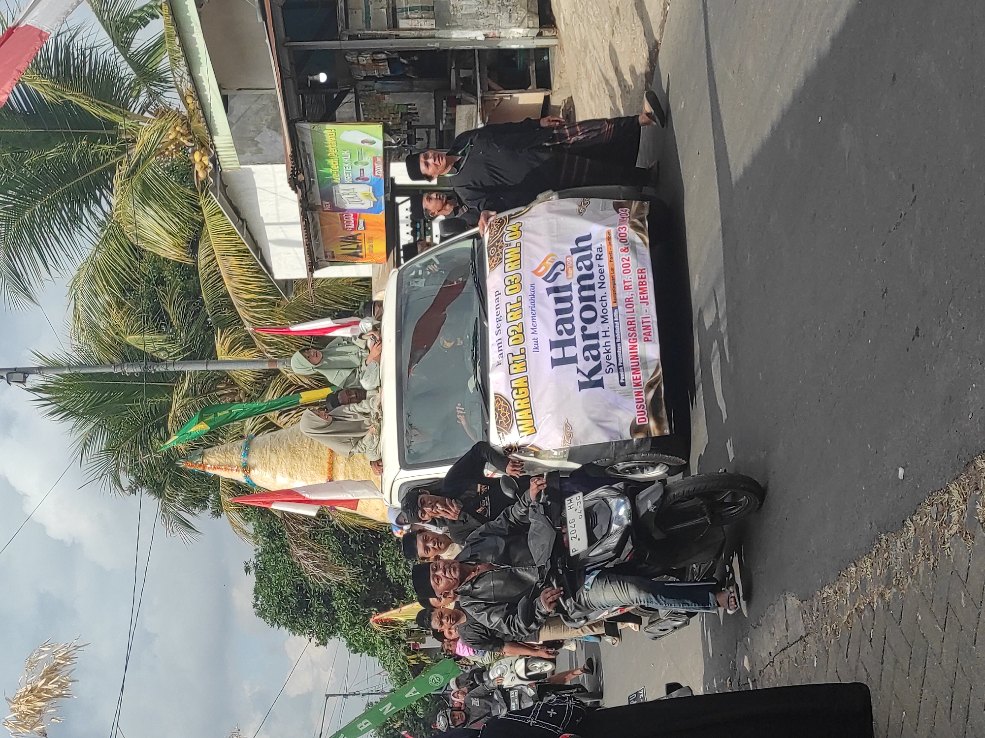 The image captures a street scene in what appears to be a procession or parade. A white van, decorated with a large banner, is the central focus. The banner announces "Haul Karomah" and lists dates and locations: "RT. 02, RT. 03 RW. 04", "Dusun Kemuningsari Lor, RT. 002 & 003", and "Panti - Jember". Several people are present. In the foreground, a man is riding a black motorcycle, looking towards the right. He is wearing a black shirt, blue jeans, and a black cap. Behind him, several other people are walking and riding motorcycles. The van has people on top, some of whom are wearing religious attire. Flags, including Indonesian flags and green flags, are visible around the van and in the background. Coconut trees and buildings line the street, suggesting a tropical or subtropical location. The sky is partly cloudy. The overall atmosphere suggests a community event or celebration.